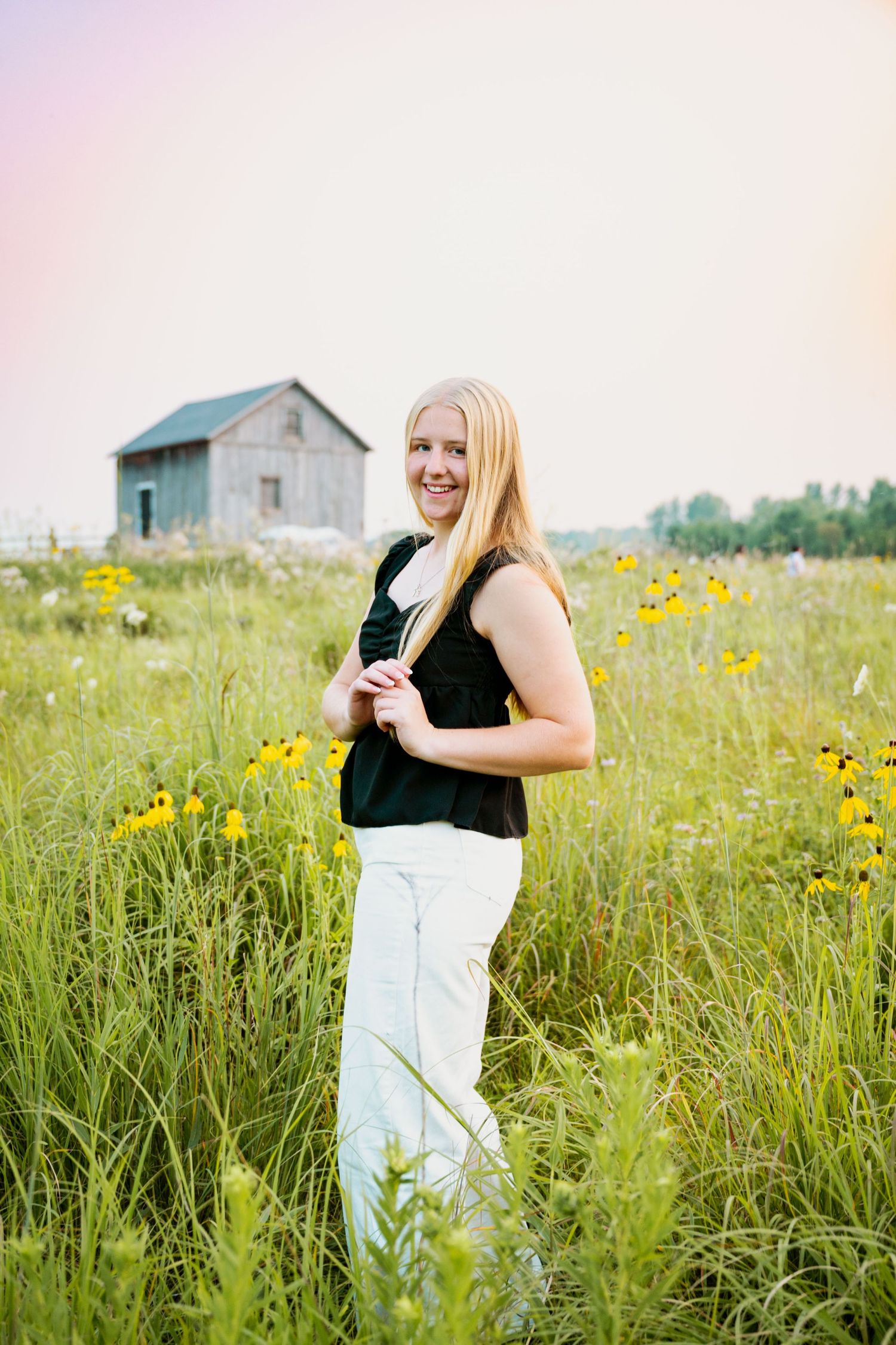 A person stands in a meadow with yellow wildflowers and tall grass, with a rustic barn in the background at sunset.