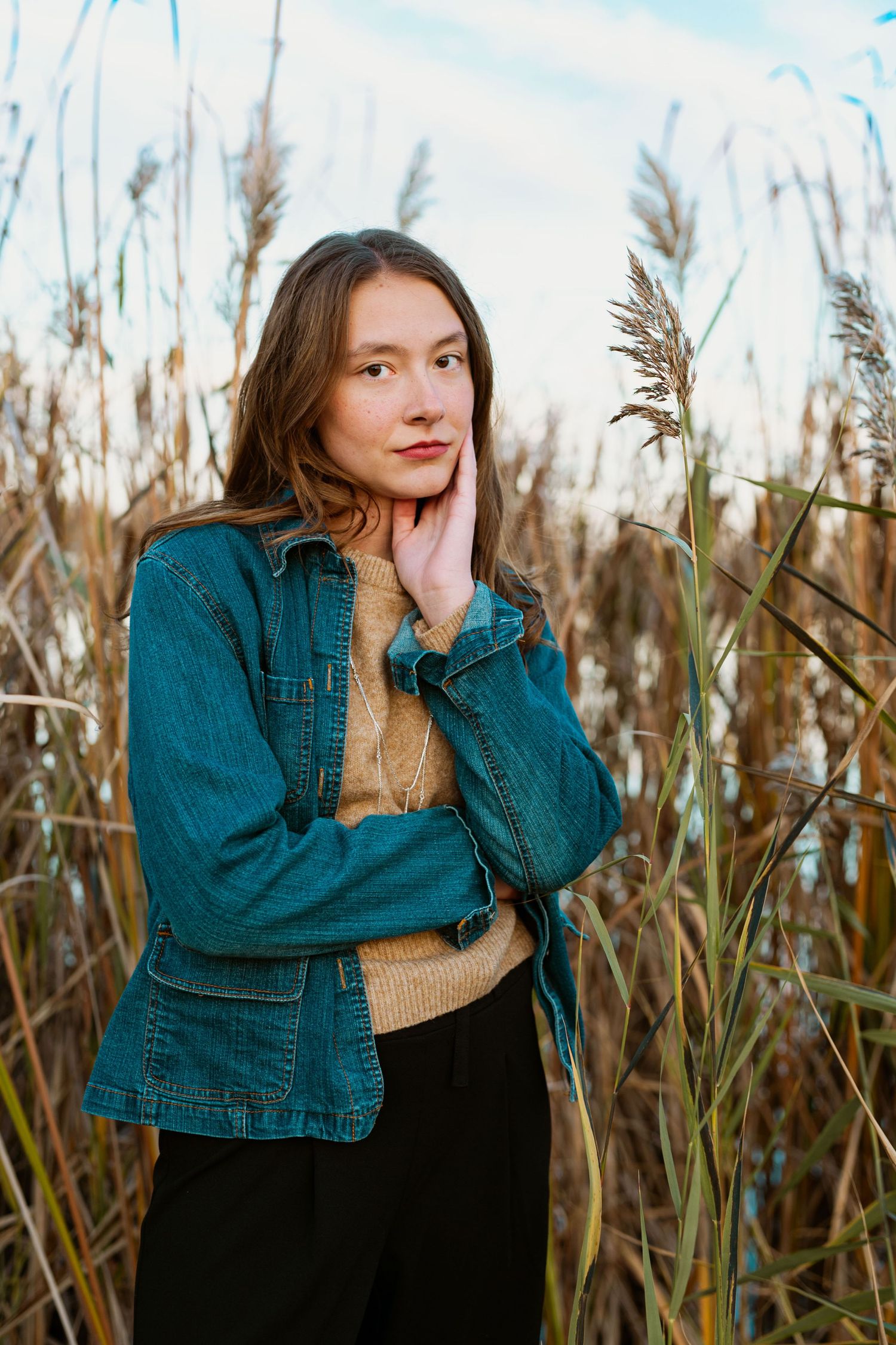 A portrait in tall grass features someone wearing a teal denim jacket and tan sweater.