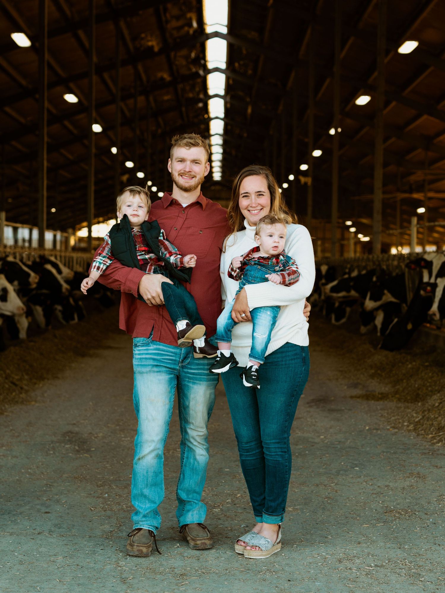 Two people in casual attire stand together holding cameras in a rustic barn setting with cattle in the background.