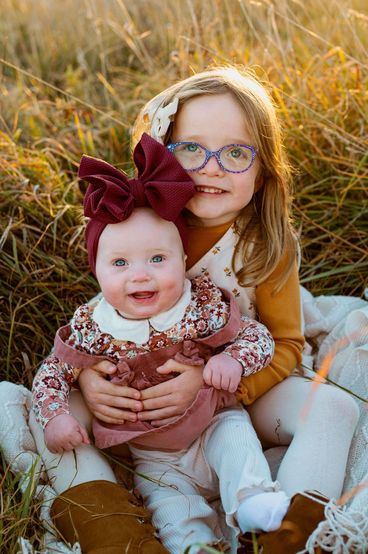 Two siblings in matching floral outfits sit together in golden sunlit grass during an outdoor autumn photoshoot.