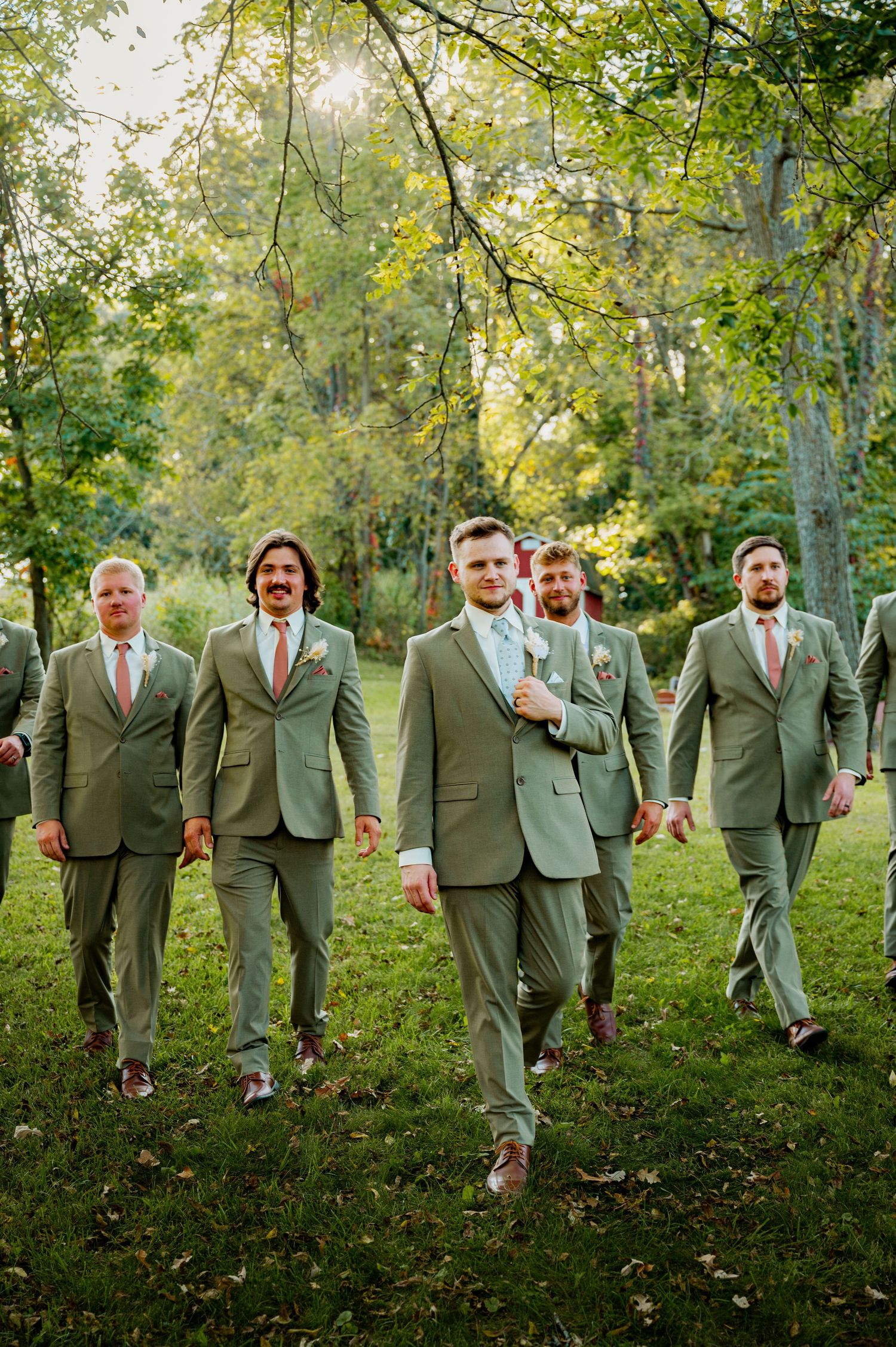 A group of groomsmen in sage green suits walk together on a grassy hillside under trees.