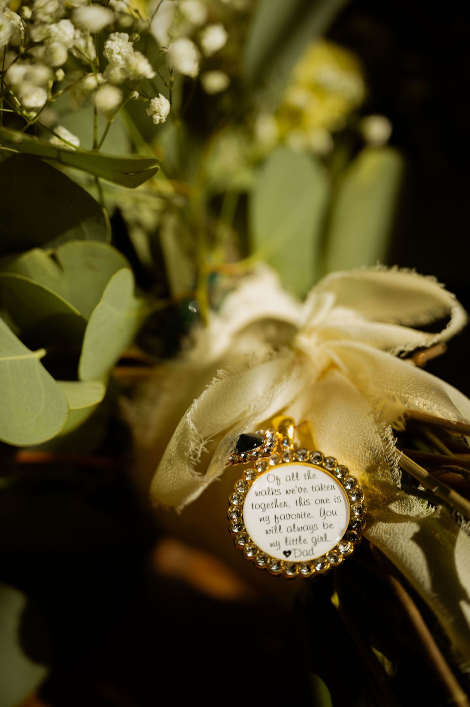 A vintage-style brooch with text rests among soft white flowers and eucalyptus leaves in warm lighting.