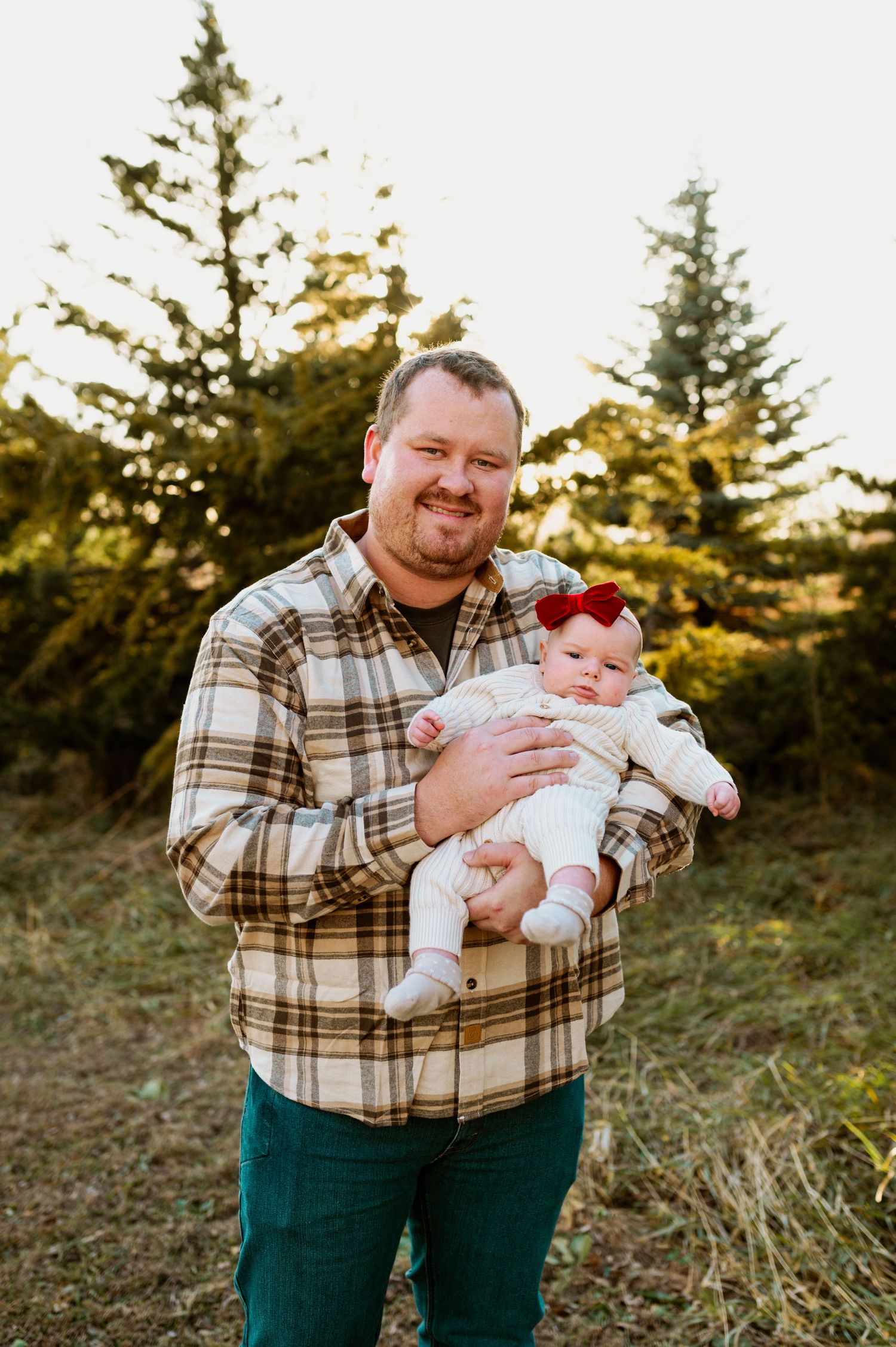 Person in plaid flannel shirt and dark pants holds baby outdoors during golden hour sunset.