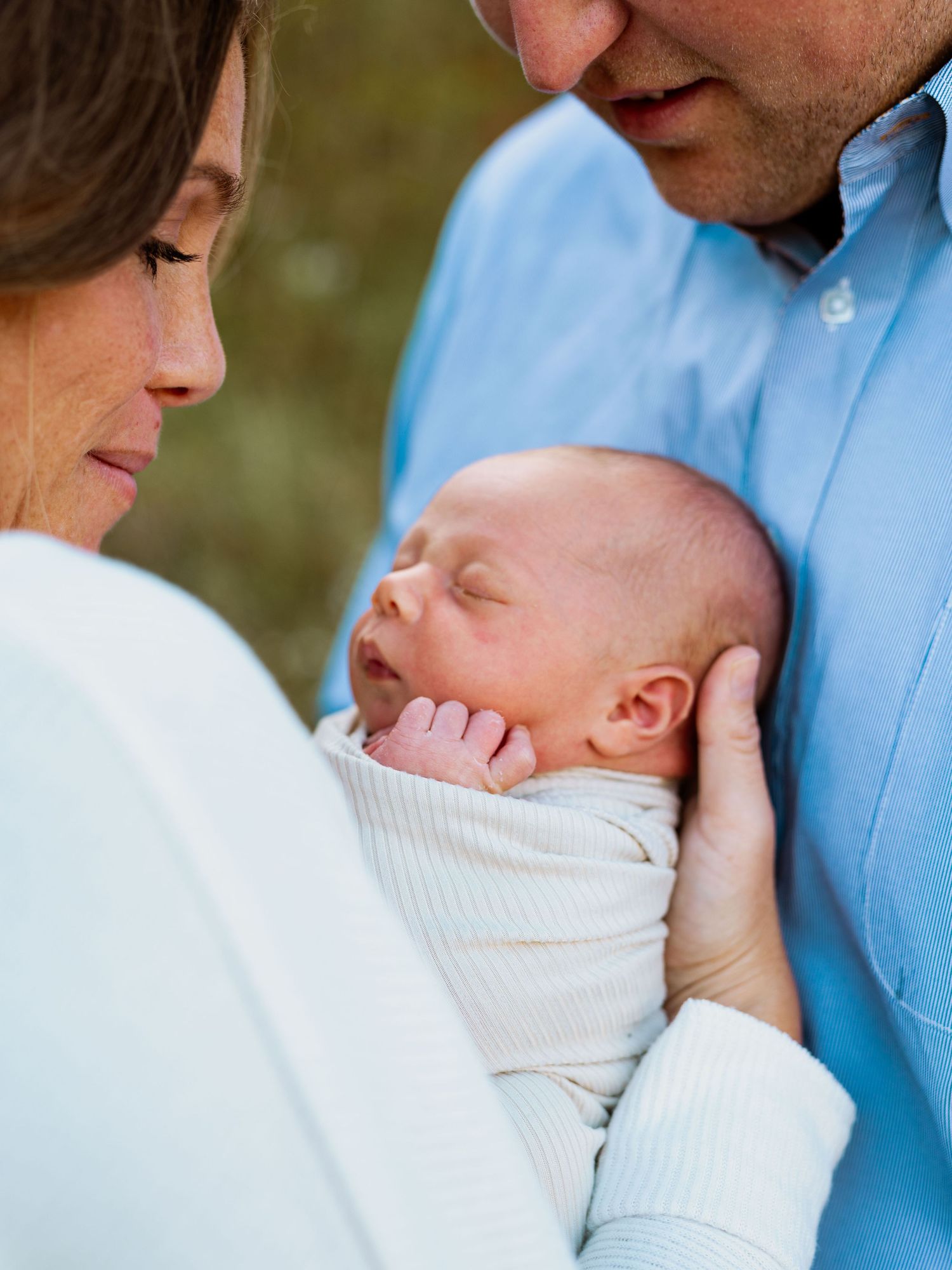 A tender moment as a parent in a blue shirt cradles and kisses a sleeping newborn baby wrapped in white.