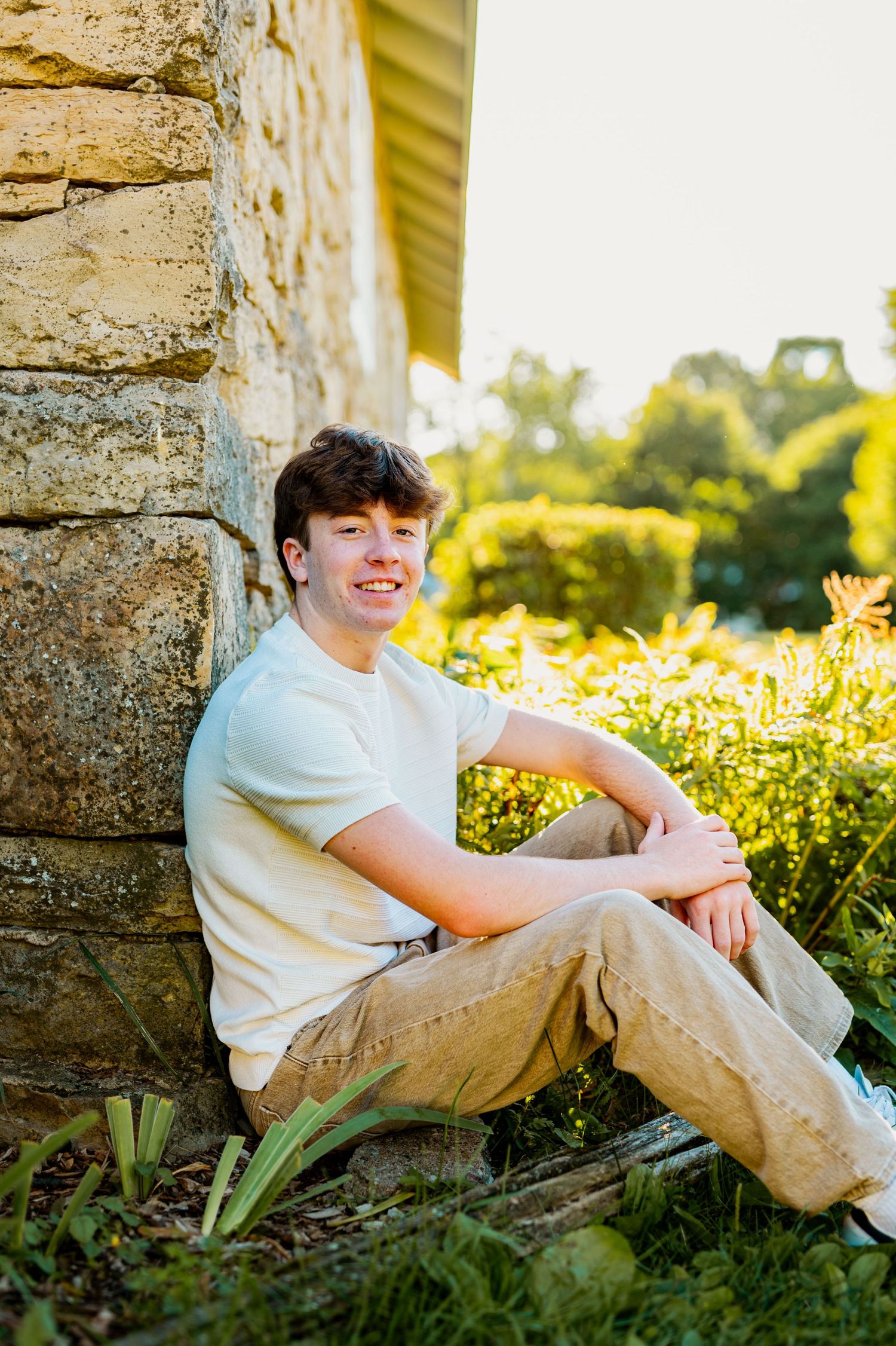 A person sits casually against a stone wall at sunset surrounded by lush greenery and golden light.