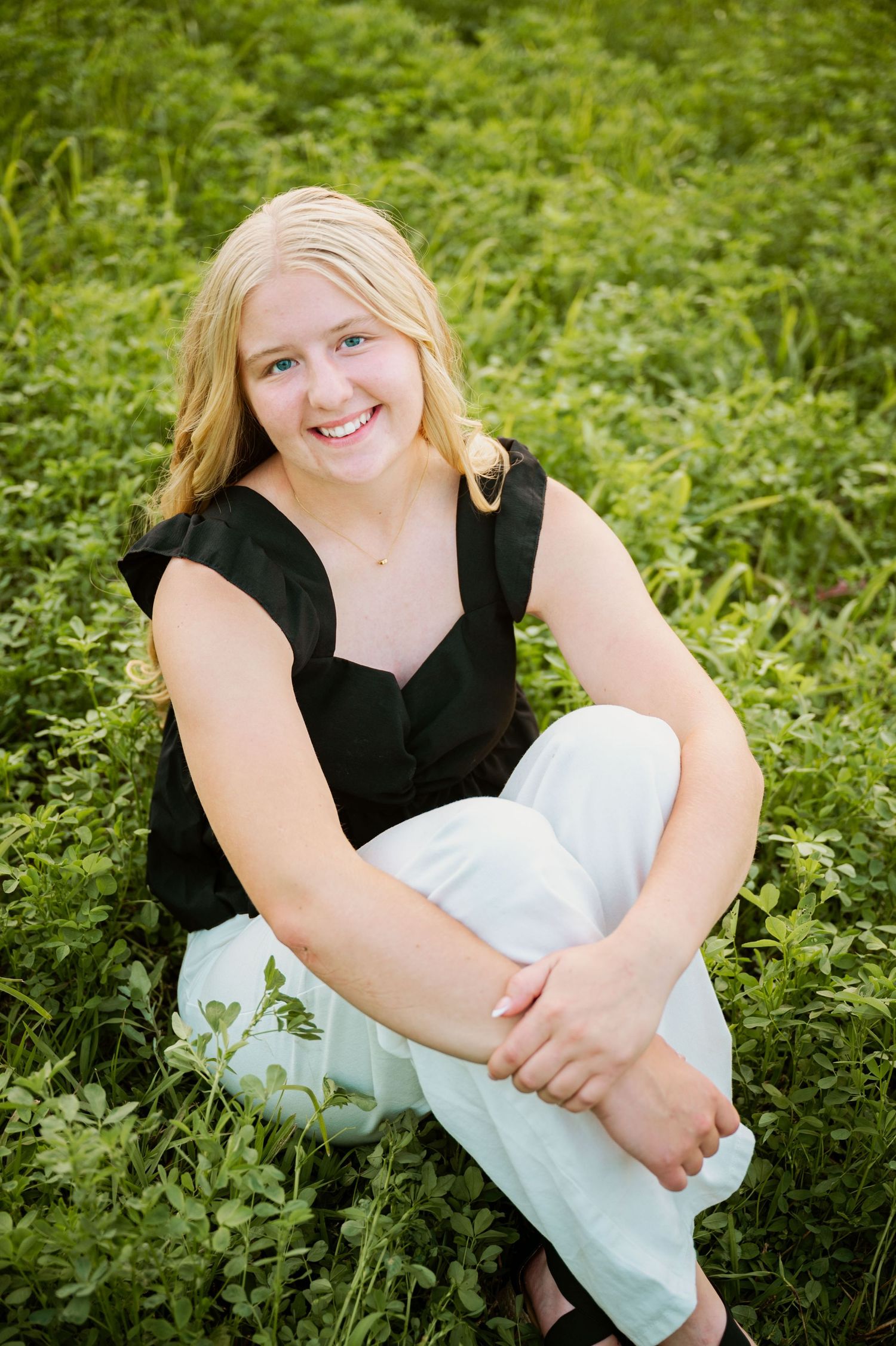 A smiling person in a black sleeveless top and light pants sits casually in green grass during an outdoor portrait session.