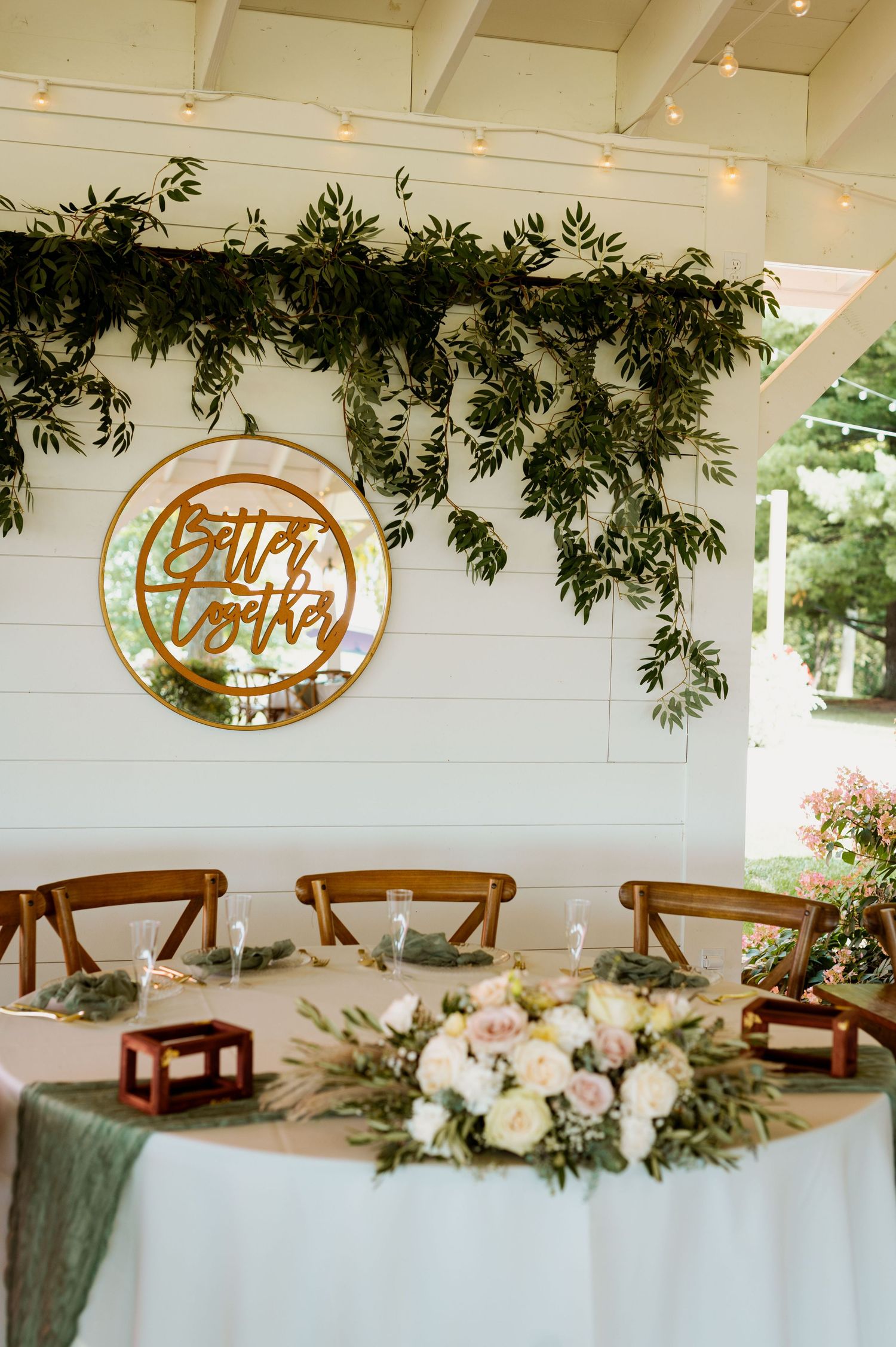 Elegant wedding reception table setup with wooden chairs and gold 'Better Together' sign under greenery and string lights.