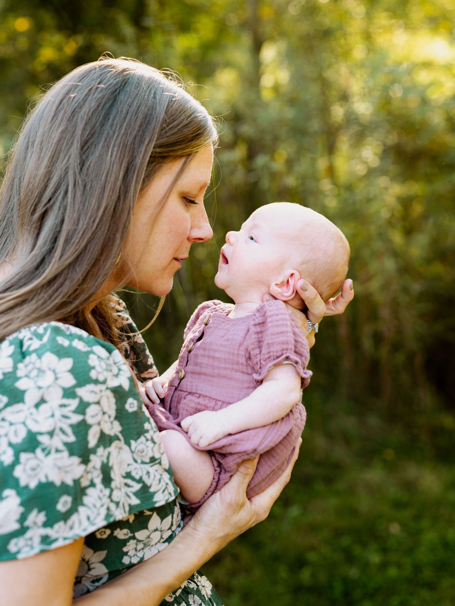 A woman in a floral dress shares tender moments with a baby in pink against a sunlit outdoor background.