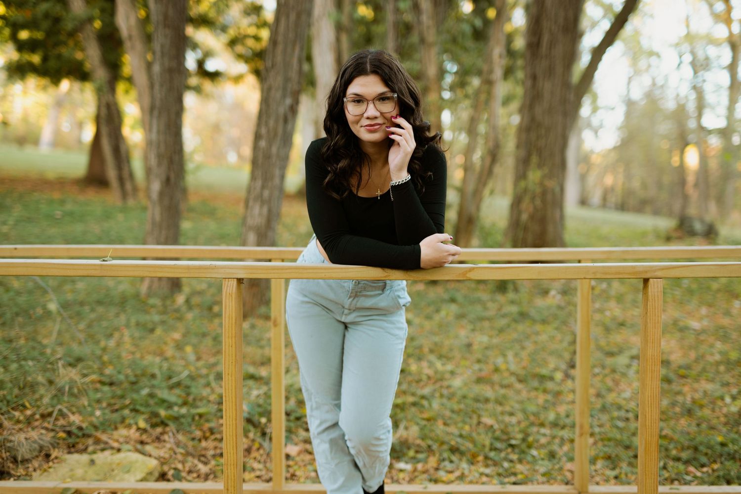 A casual portrait of someone in light jeans and black top leaning against a wooden railing in a park setting.