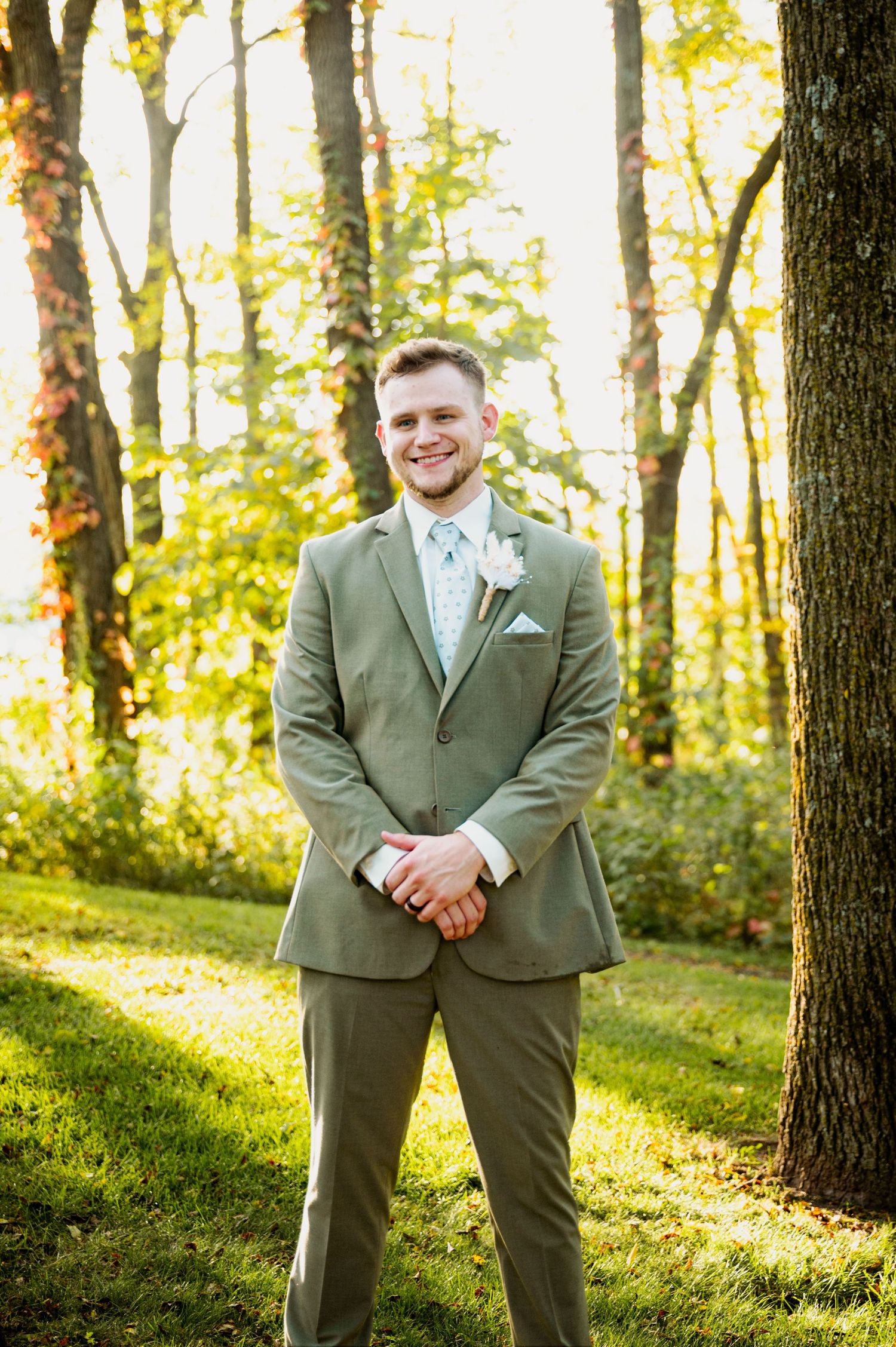 Groomsman in sage green suit poses confidently in a sunlit forest setting.