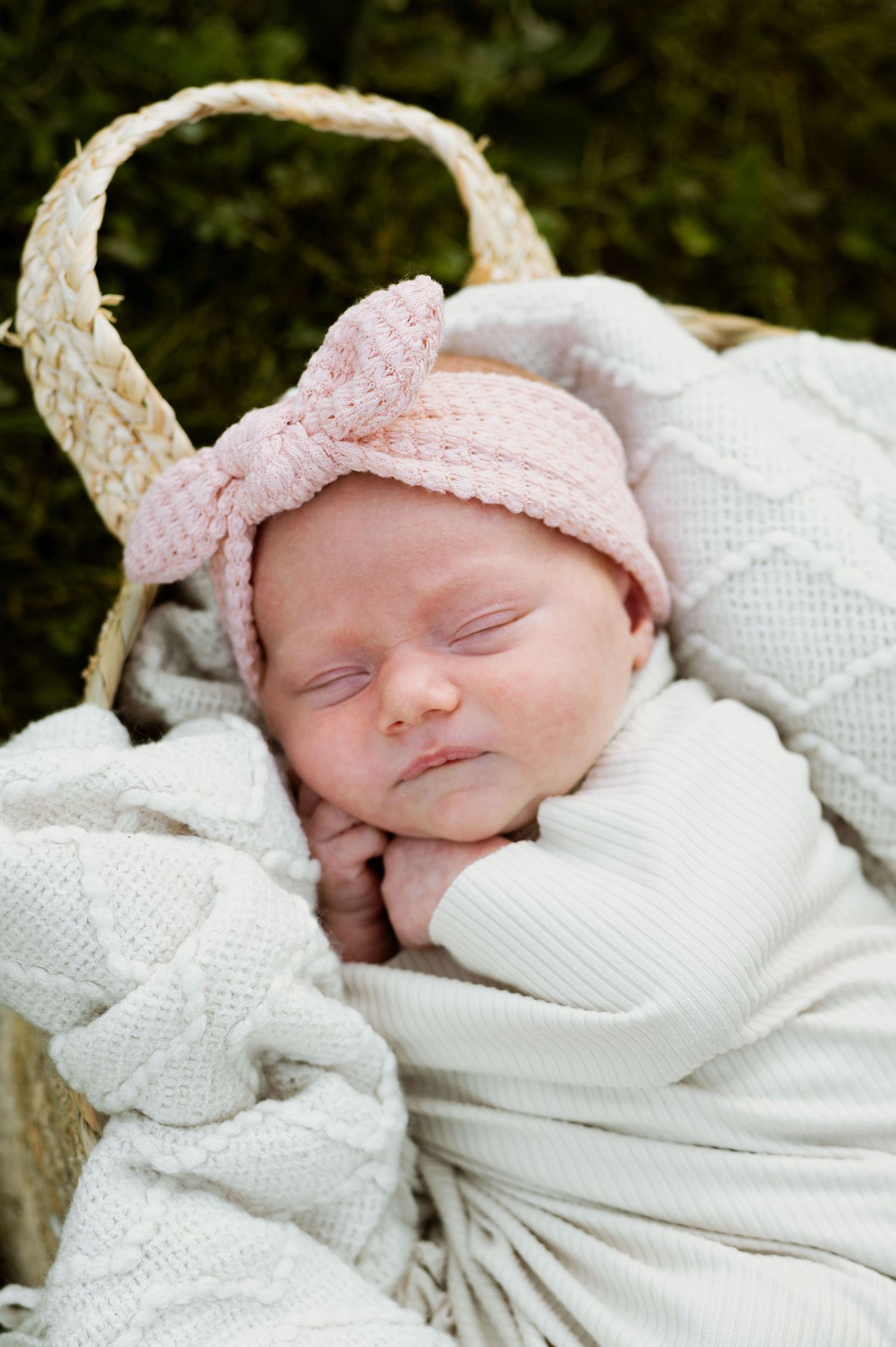 A sleeping newborn baby wearing a white knit outfit and pink bow headband rests peacefully on white blankets.