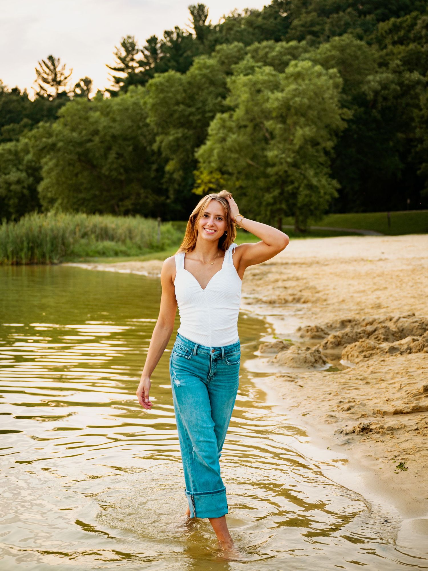 A person in a white tank top and teal pants stands ankle-deep in water along a scenic beach with lush trees in the background.