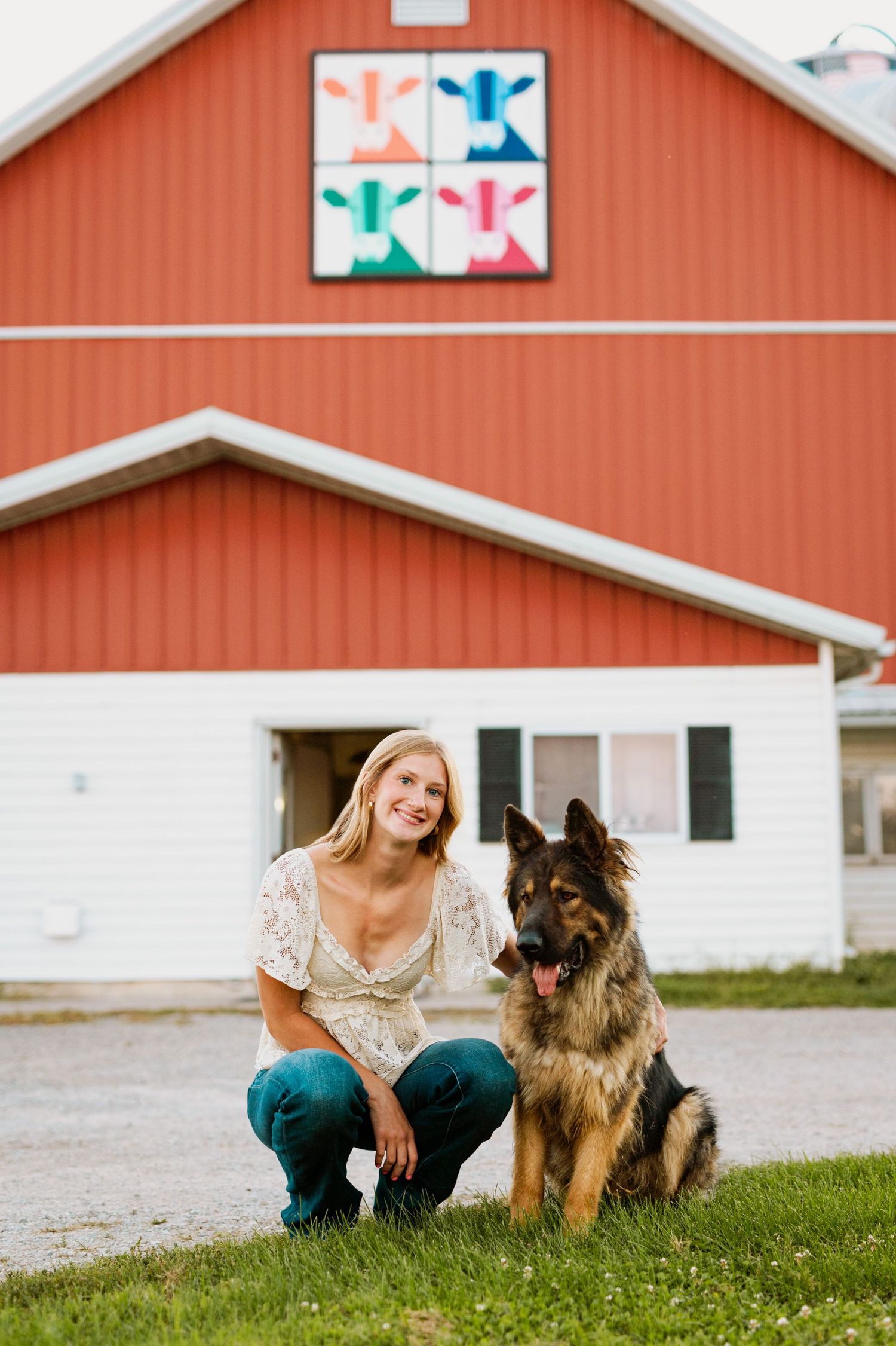 A German Shepherd sits beside someone on grass in front of a red barn with a decorative quilt pattern on its exterior.