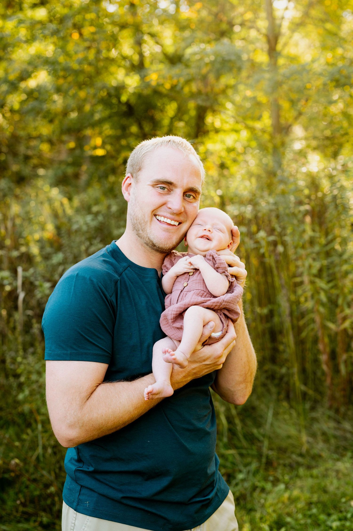 A tender outdoor portrait showing a parent in a navy shirt holding a small baby against a sunlit forest backdrop.