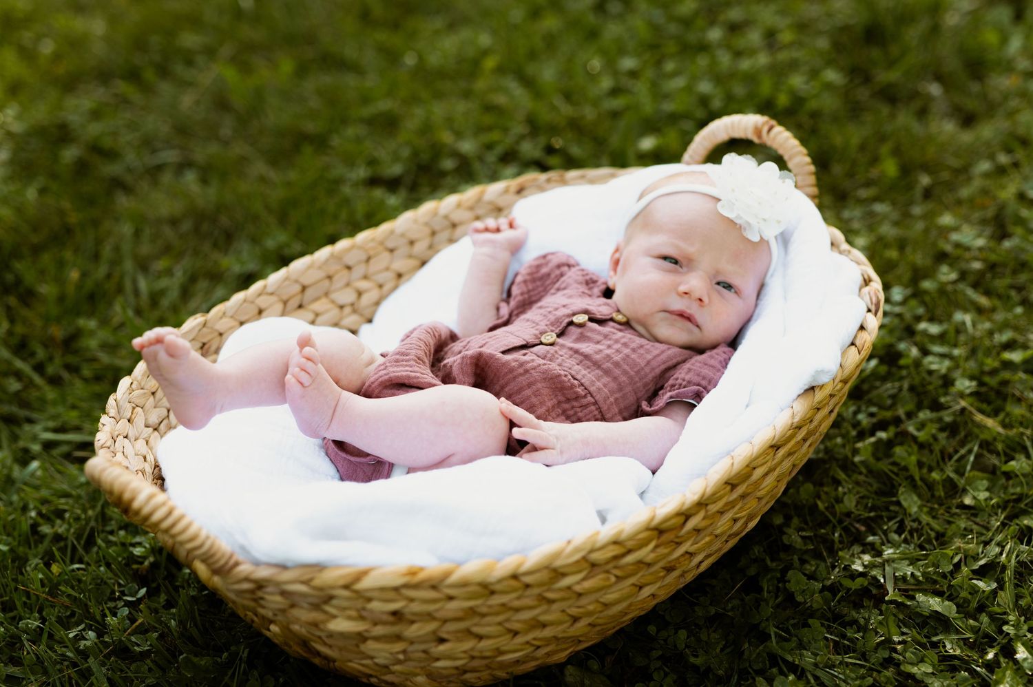A sleeping baby lies peacefully in a woven basket outdoors on green grass in natural lighting.