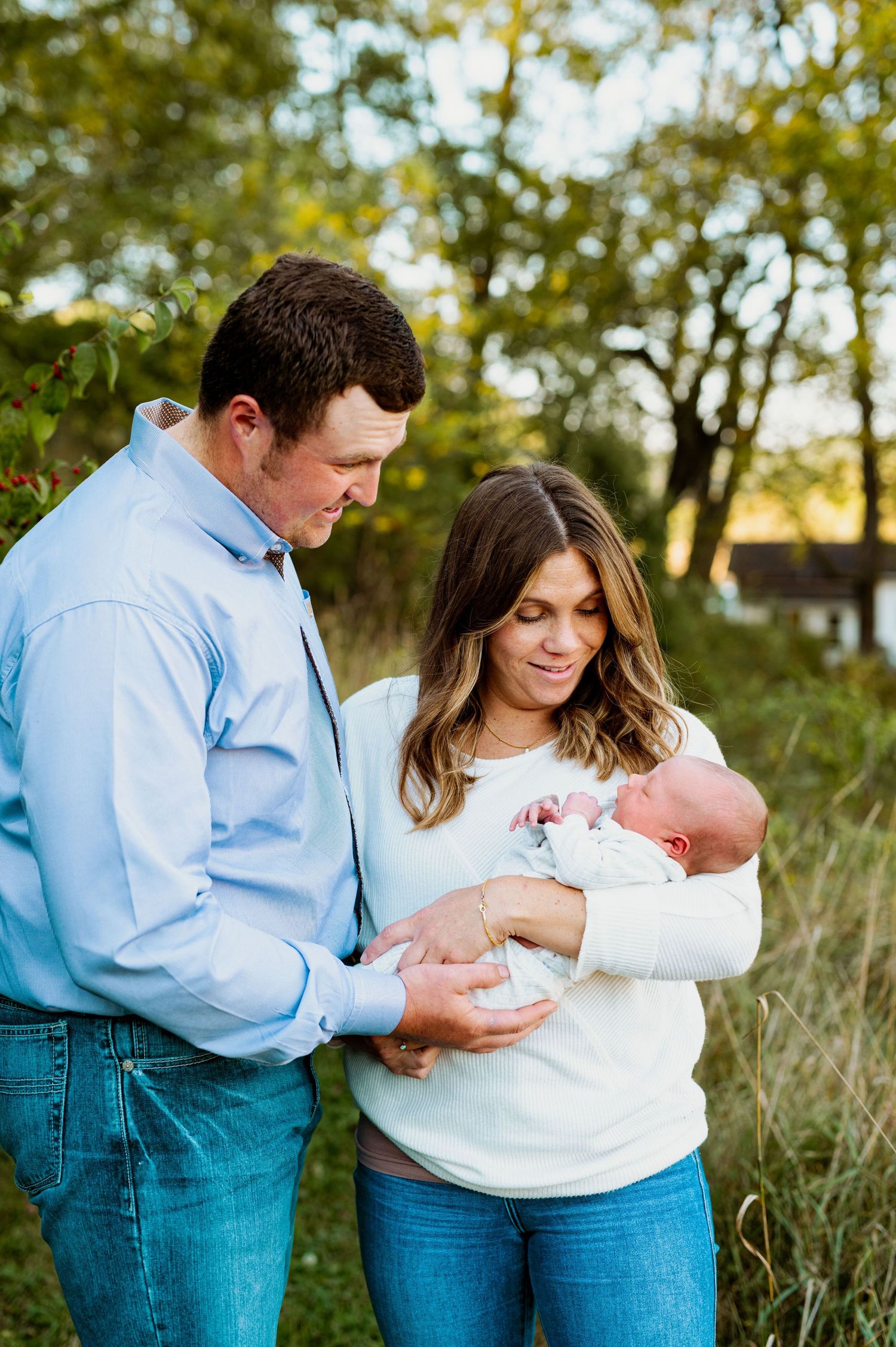 Parents in casual attire share intimate moments with their newborn baby outdoors during golden hour.
