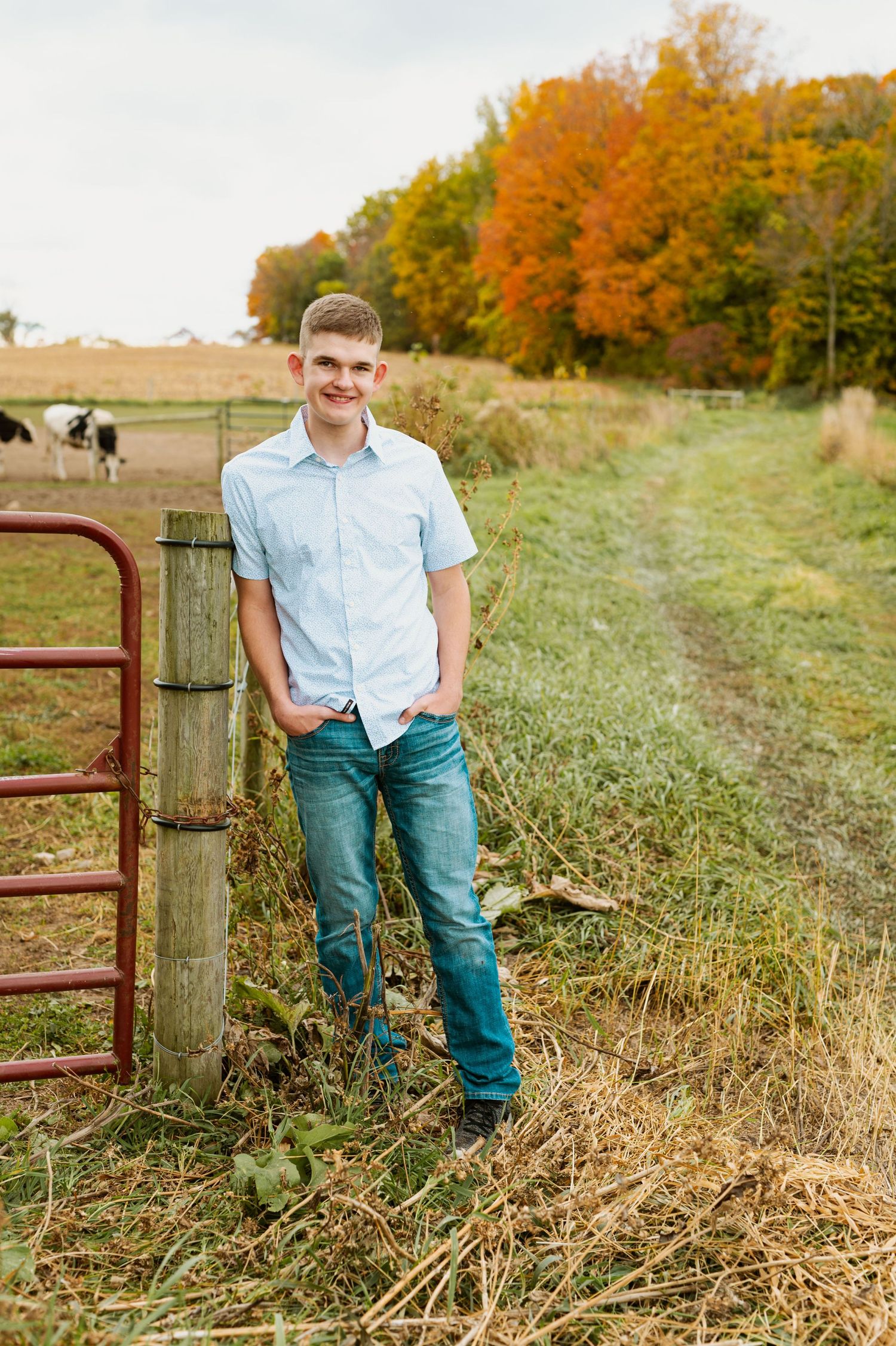 Casual outdoor portrait by rustic farm gate with fall foliage and rolling hills in background.