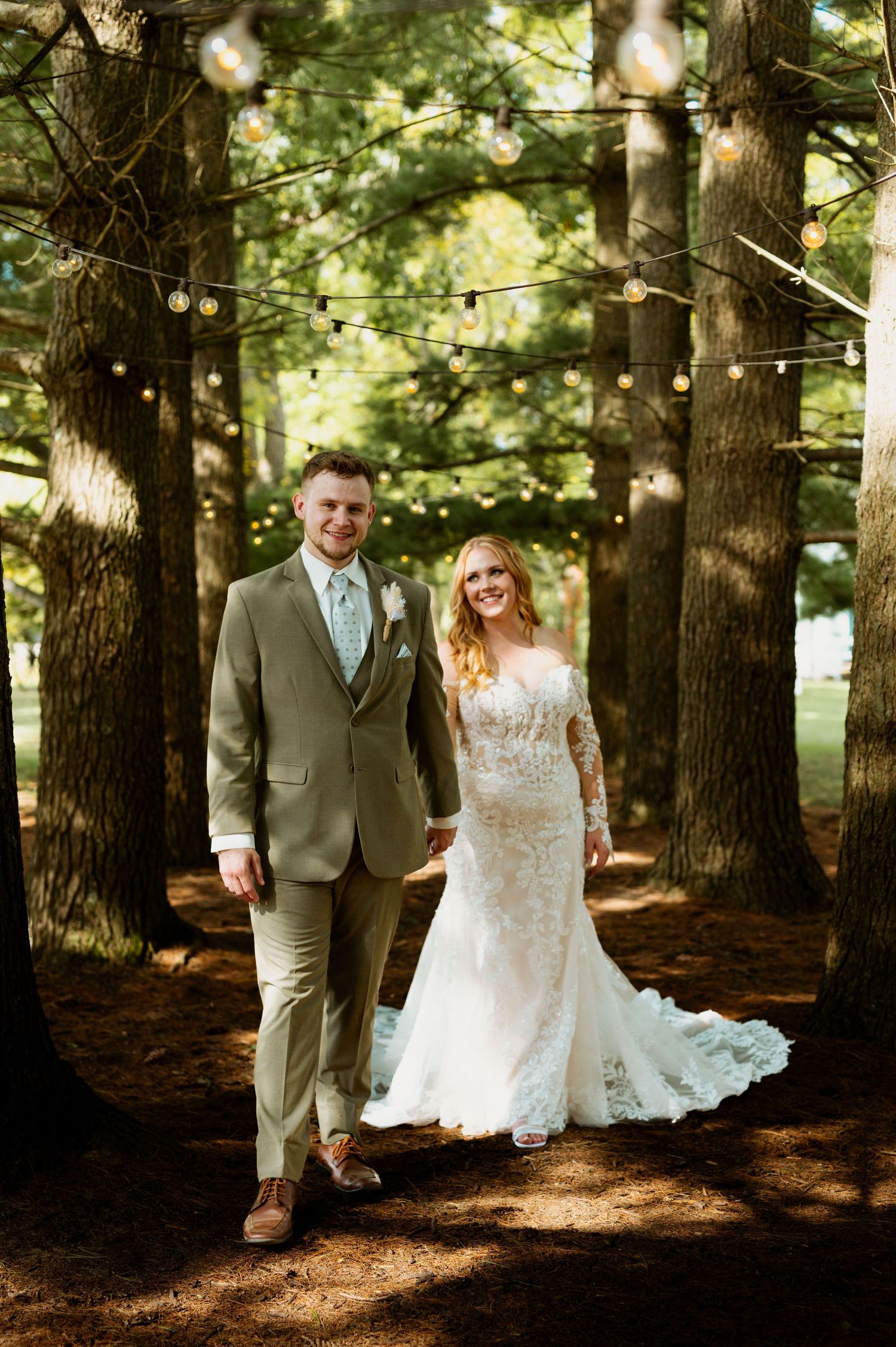 Newlyweds in a lace gown and tan suit share joyful moments while walking through a pine forest.