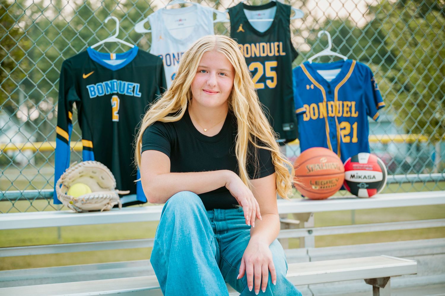 Student athlete wearing black shirt and blue jeans sits on bleachers with sports jerseys and balls displayed behind them.
