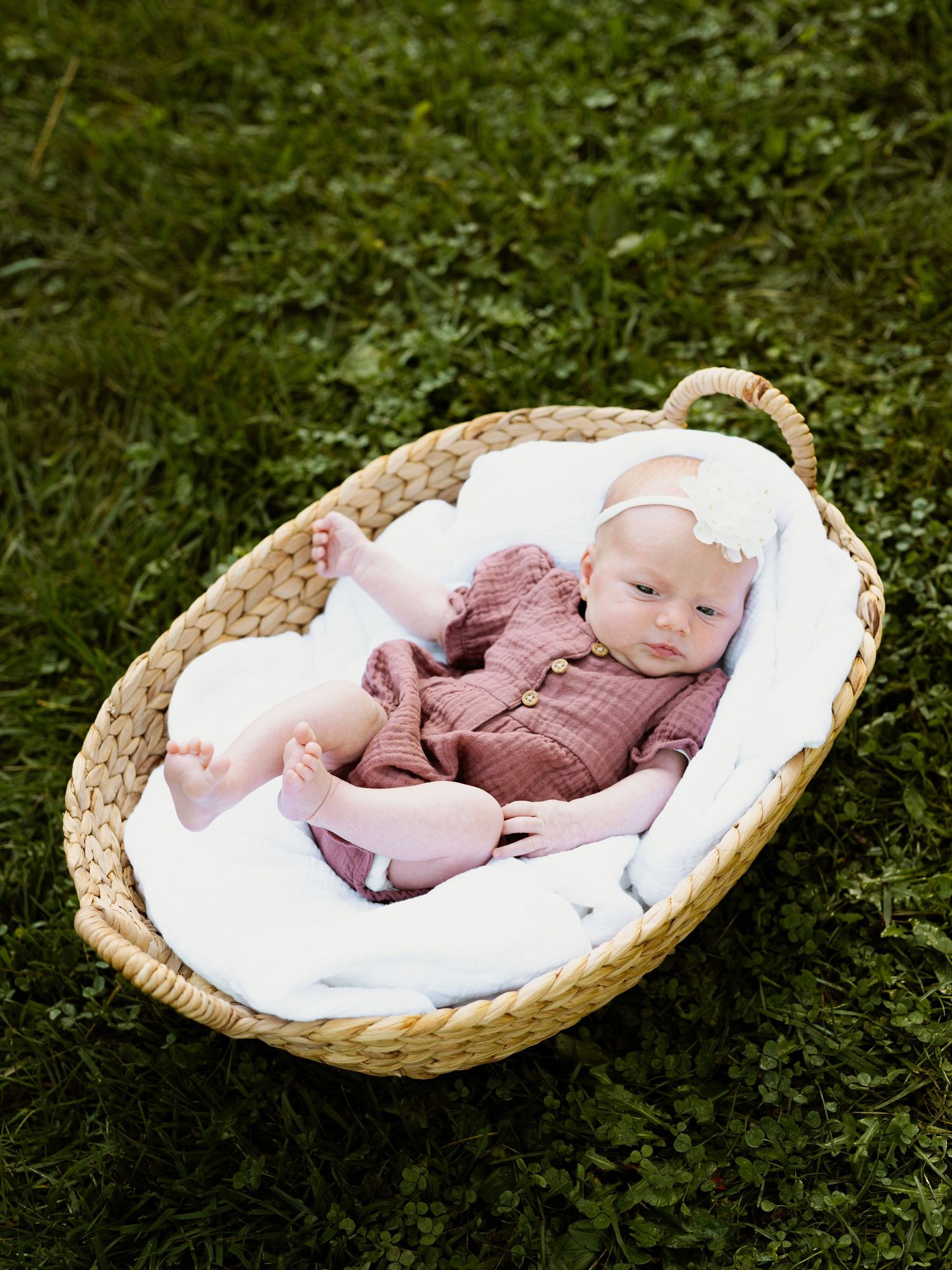 A newborn baby sleeps peacefully in a woven basket lined with white bedding placed on green grass outdoors.