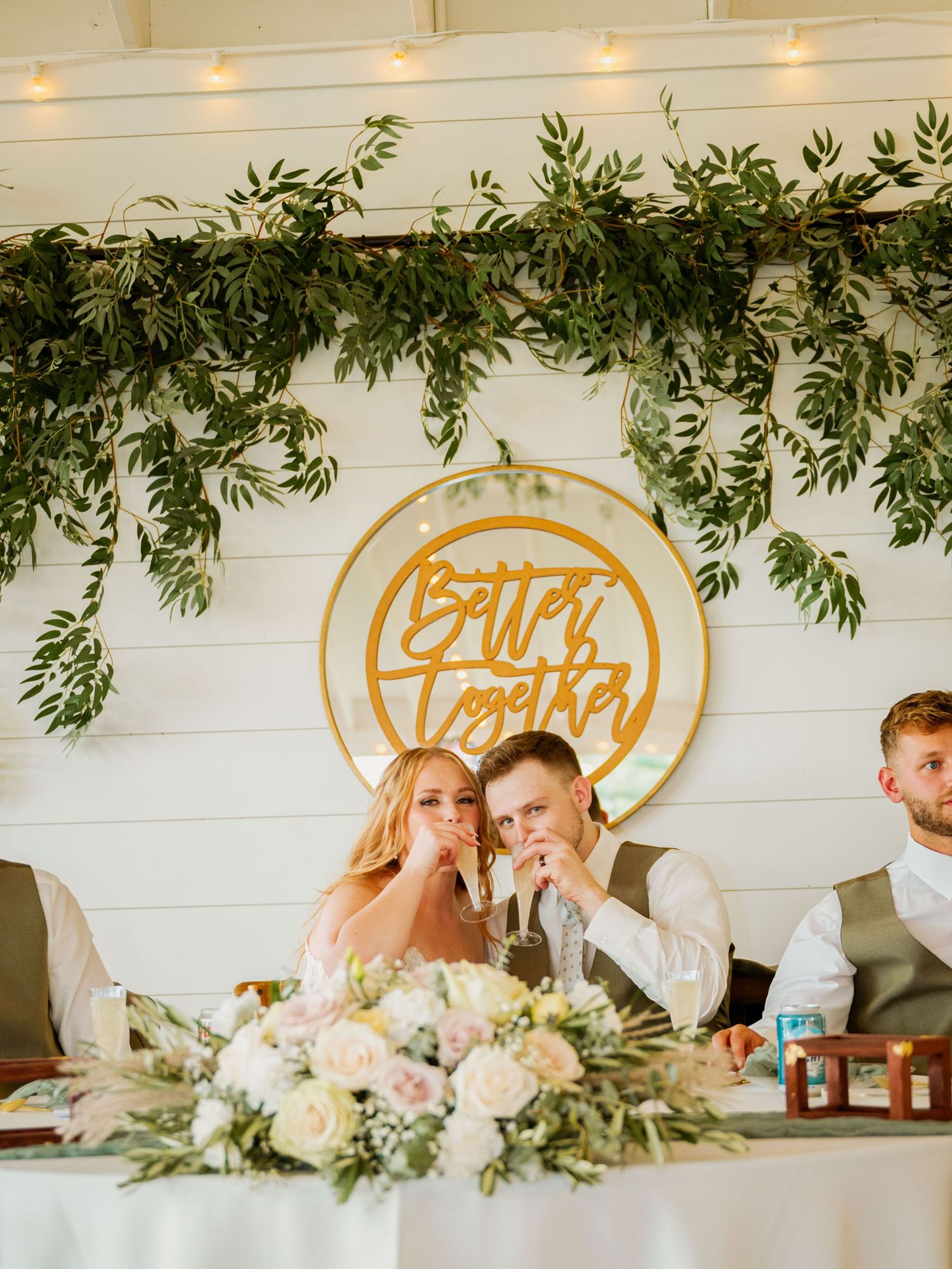 An elegant reception table features greenery decorations, string lights, and a gold circular wedding sign on white walls.
