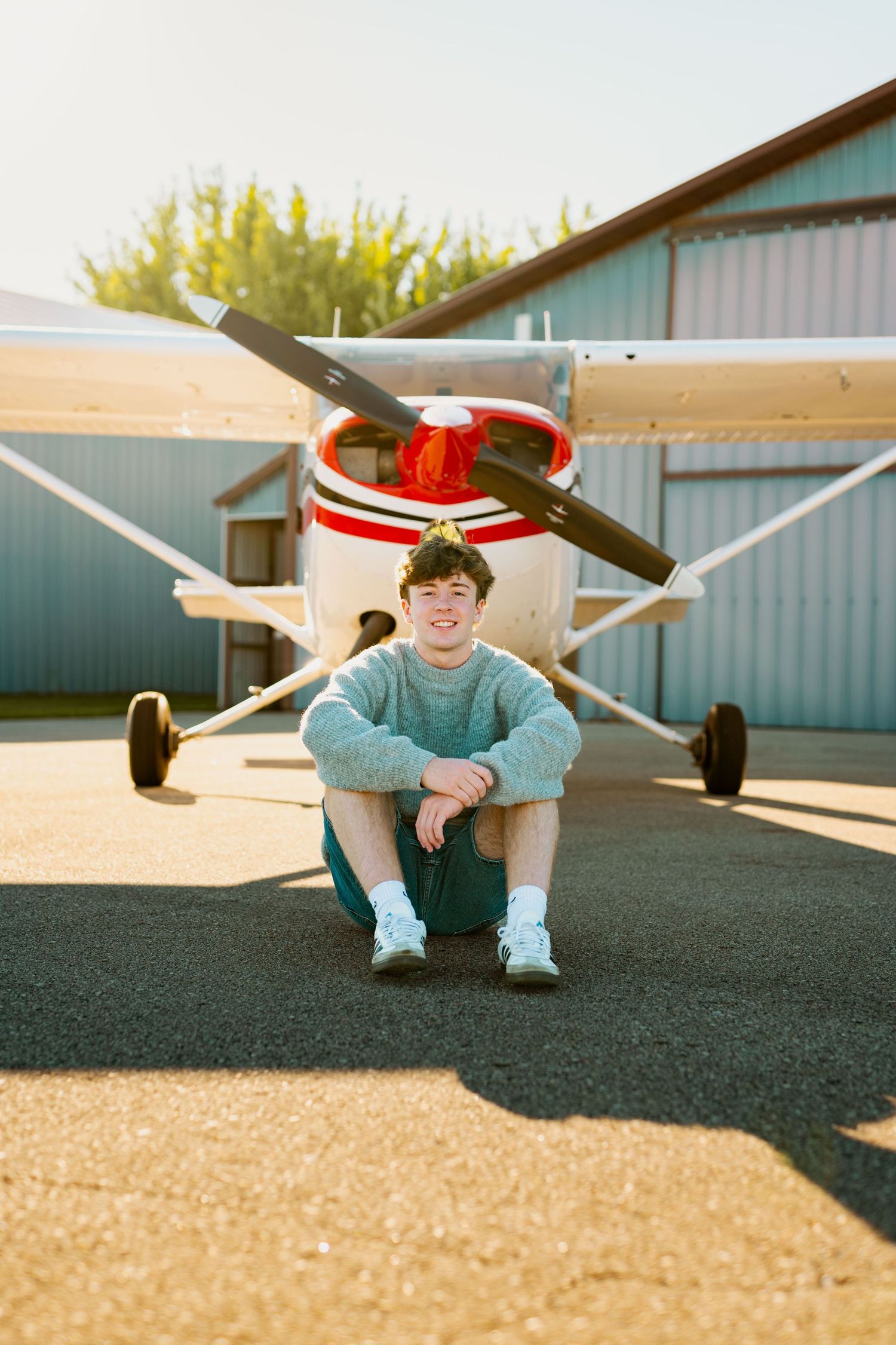 A small aircraft parked on a tarmac with someone sitting beneath the wing in a casual pose.