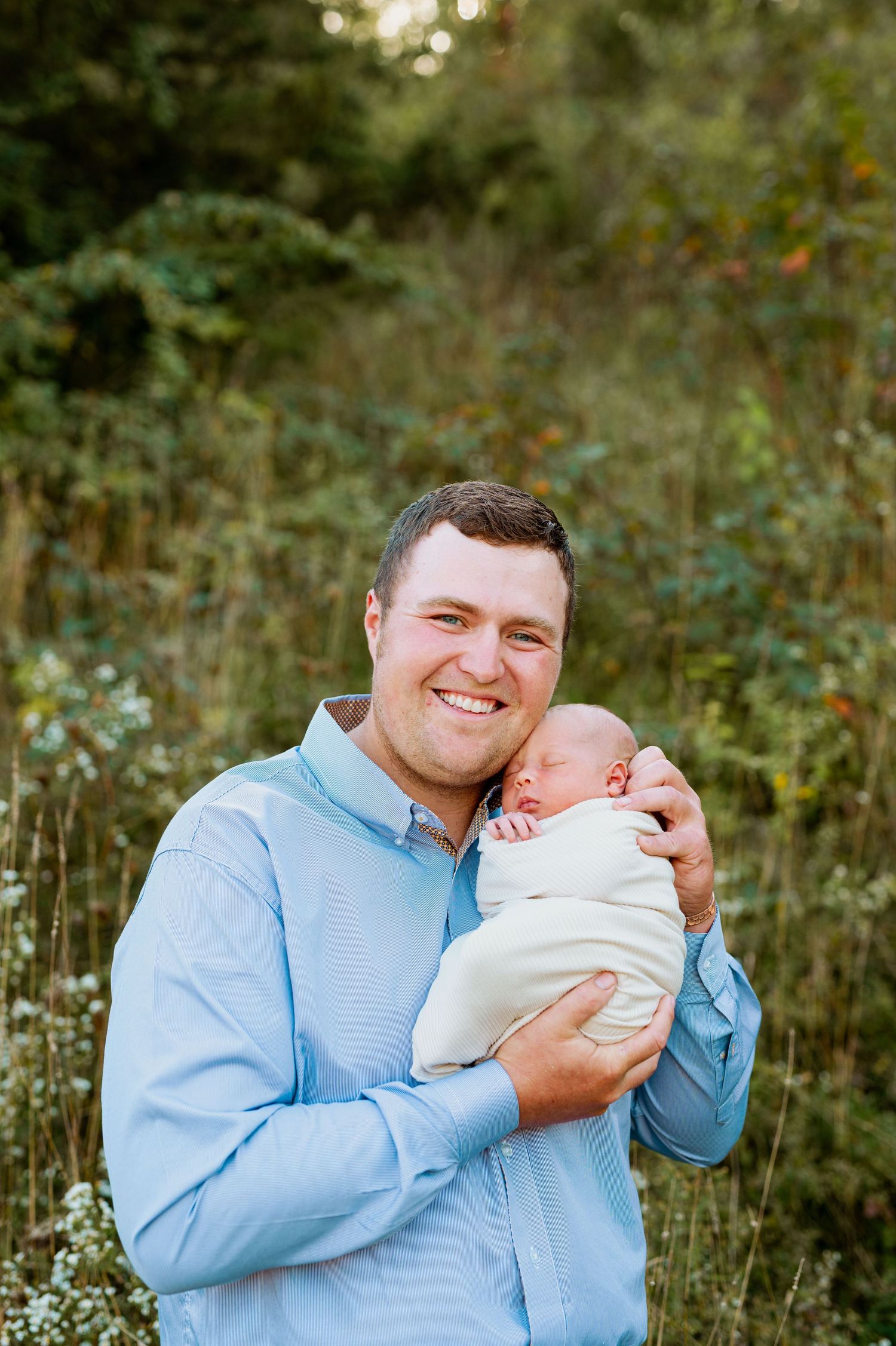 A man in a blue shirt holds and smiles at a newborn baby in a natural outdoor setting.