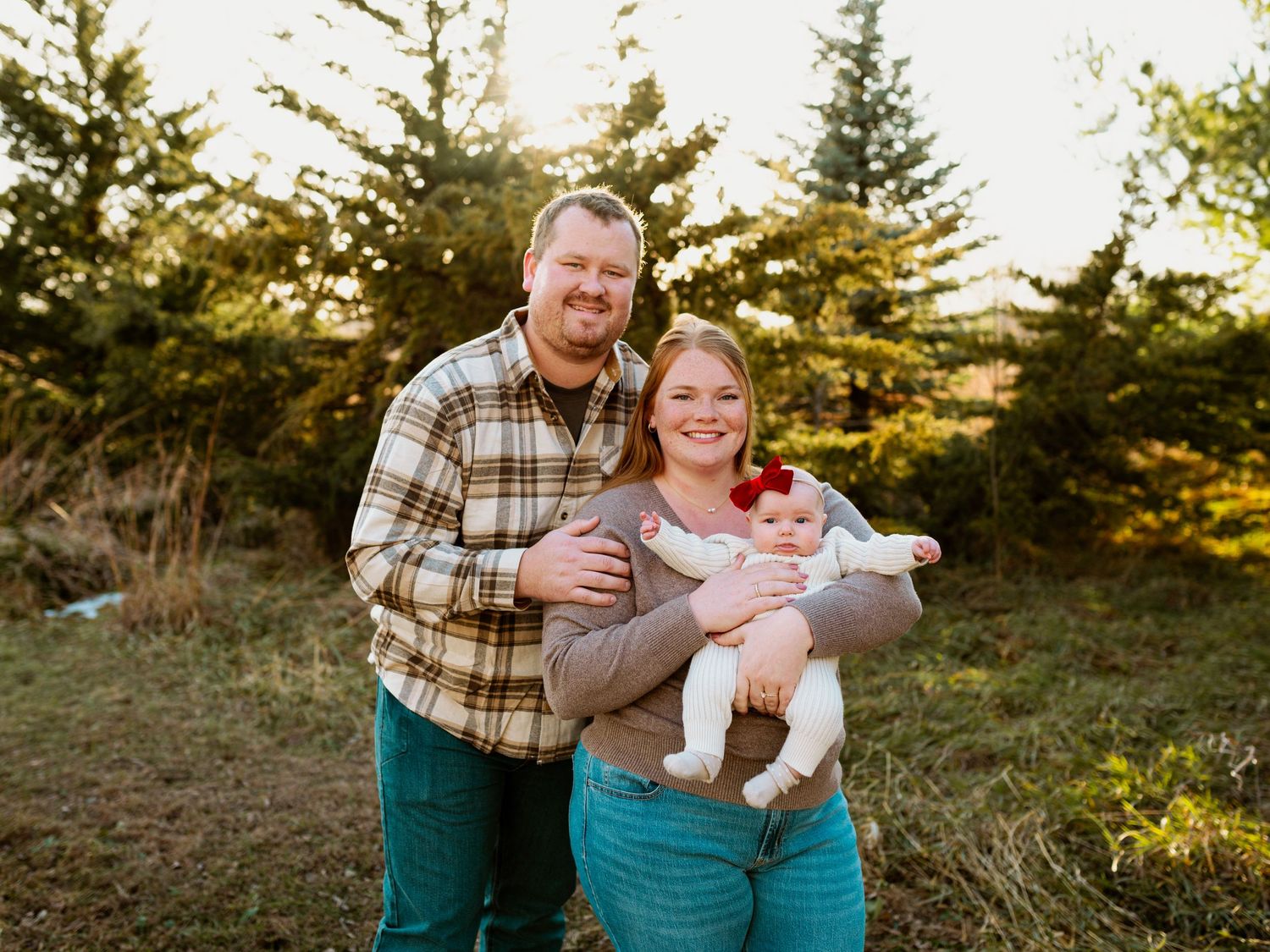 A family wearing plaid and teal sits together on a grassy hillside during golden hour sunset.