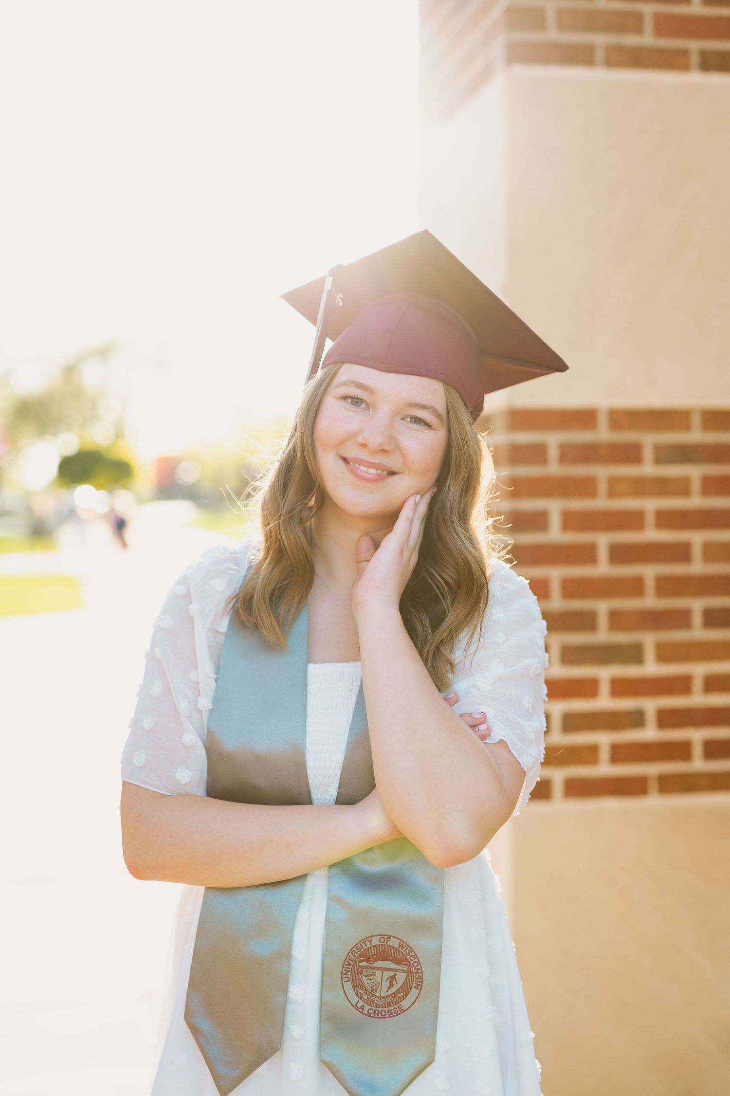 A graduate in cap and stole poses by brick column in warm evening light.