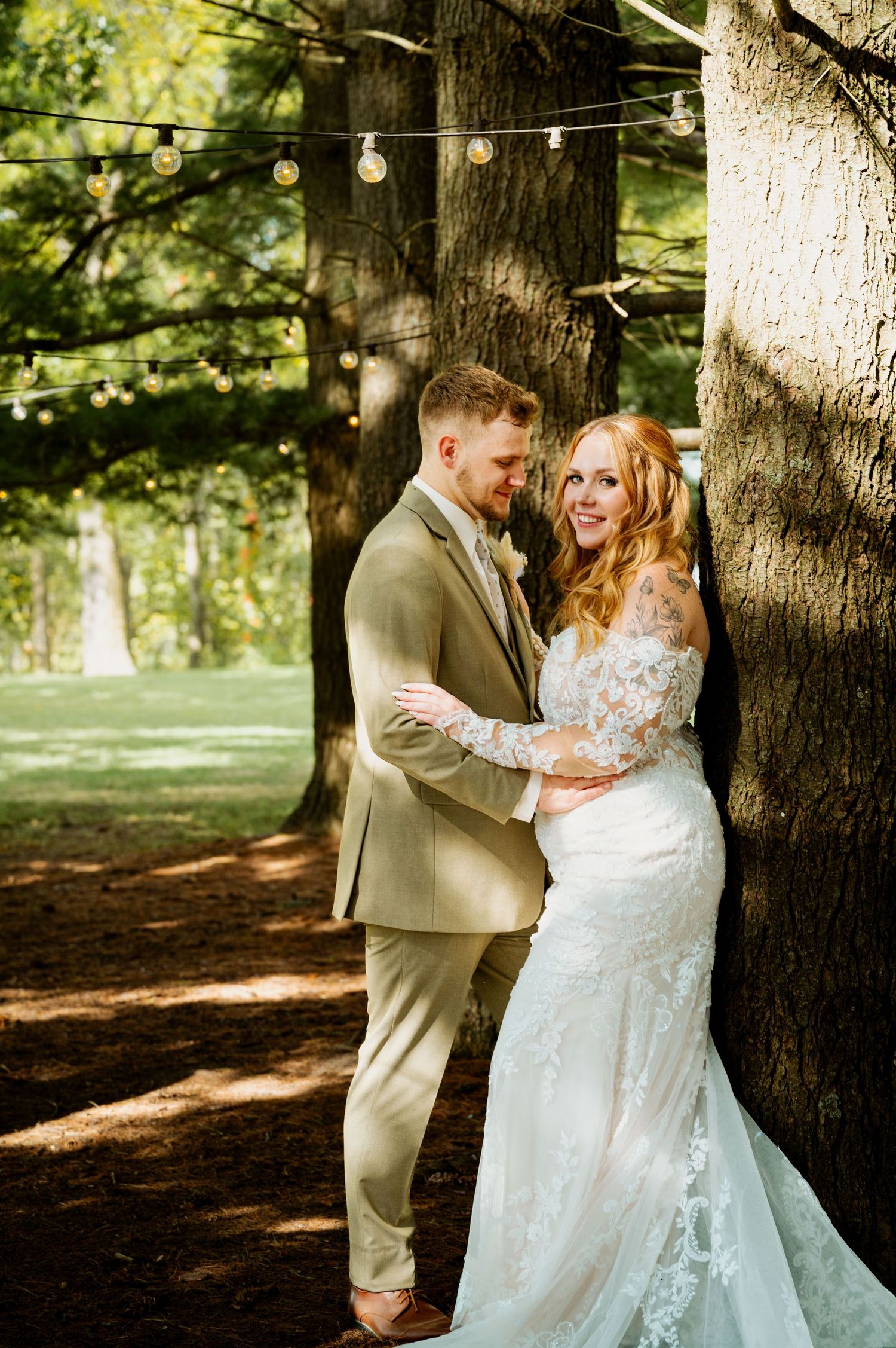 Joyful wedding couple sharing intimate moment under string lights in wooded area.