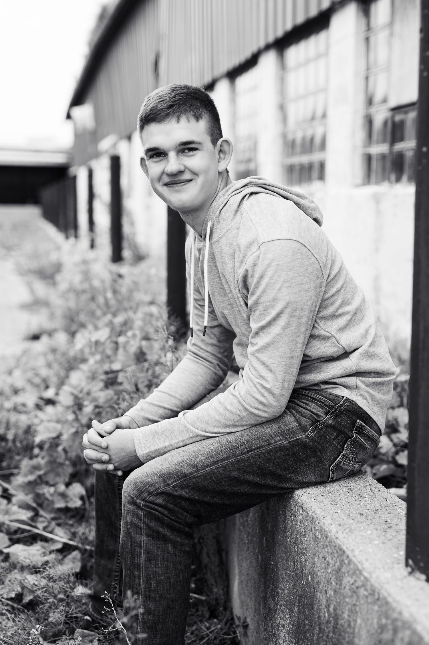 Black and white portrait of person in hoodie sitting on concrete ledge outside barn building.
