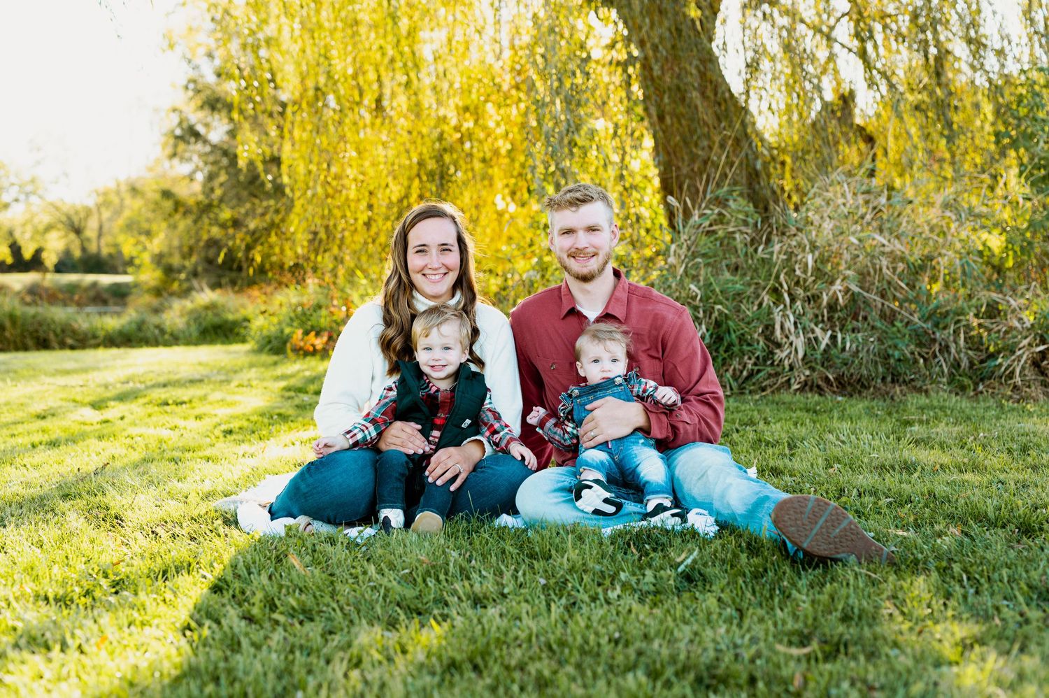 Family wearing coordinated teal and burgundy outfits sits together on grassy hill during golden autumn sunset.