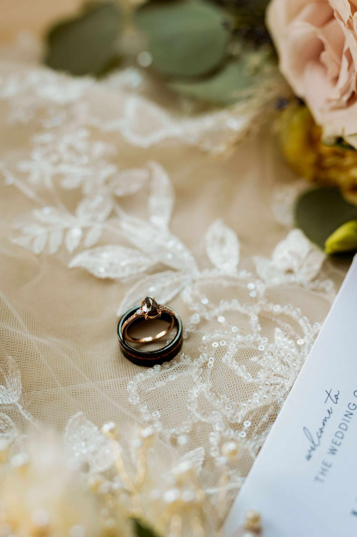 Wedding rings and invitation details rest on delicate white lace in a soft, romantic composition.