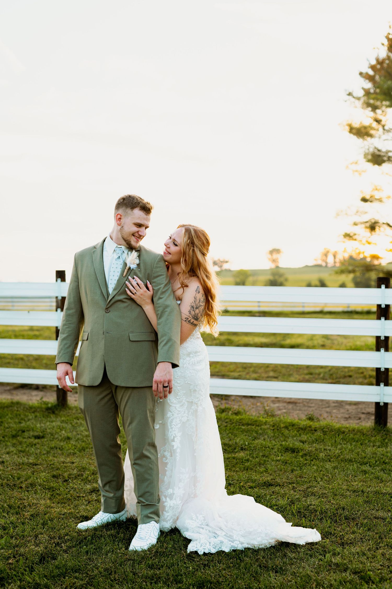 Newlyweds in elegant attire share romantic moments by white fence during golden hour.