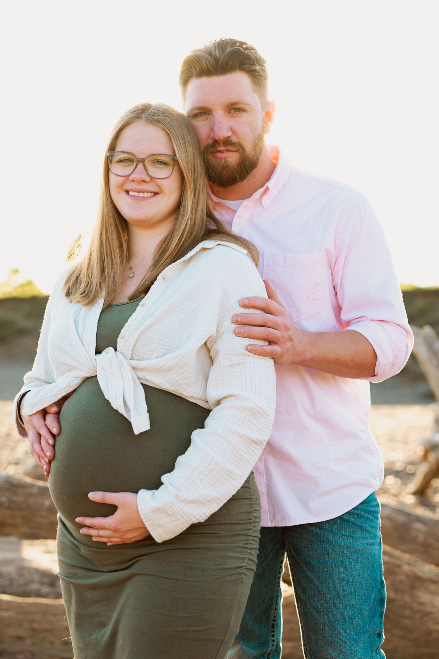 A pregnant couple poses outdoors during sunset, standing together in casual attire with warm lighting and natural scenery.