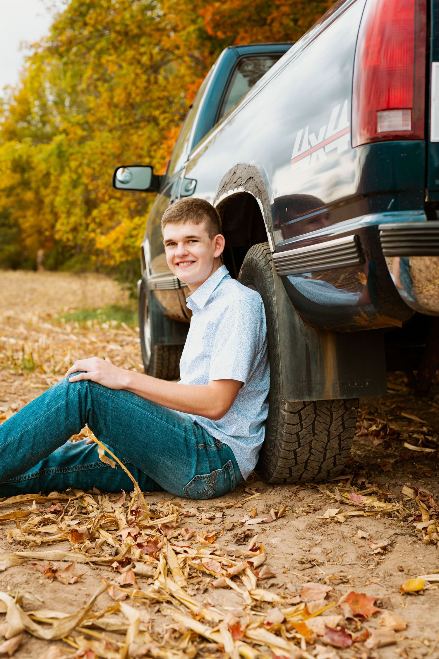 Senior portrait of student leaning against black pickup truck with fallen leaves on ground.