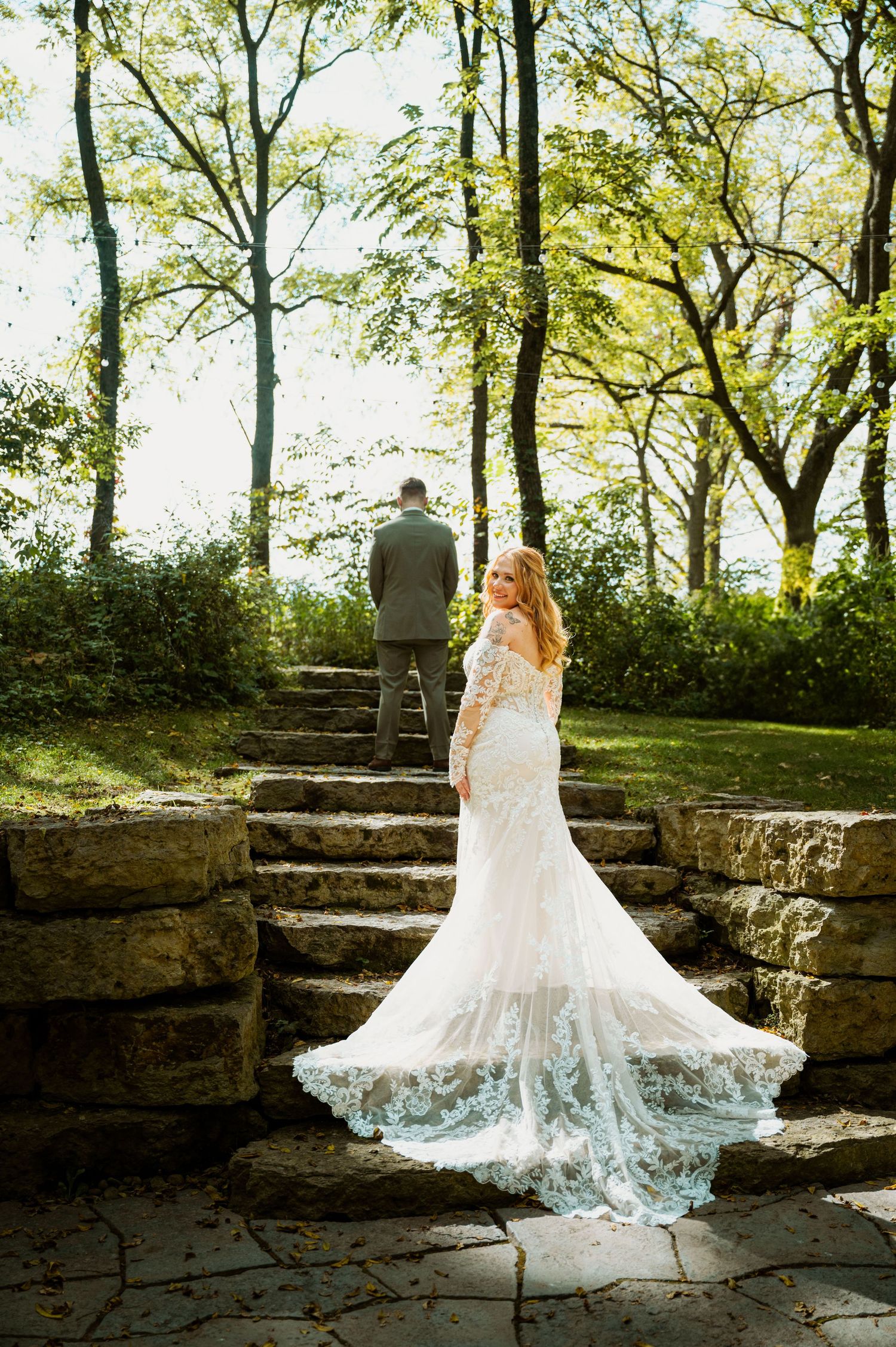 A bride in a flowing lace wedding gown poses on stone steps surrounded by lush greenery in an outdoor setting.