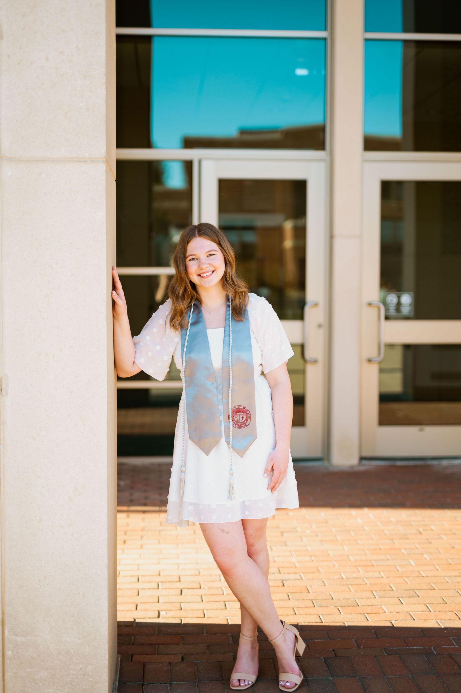 A graduate poses in white dress and stole against modern building entrance.
