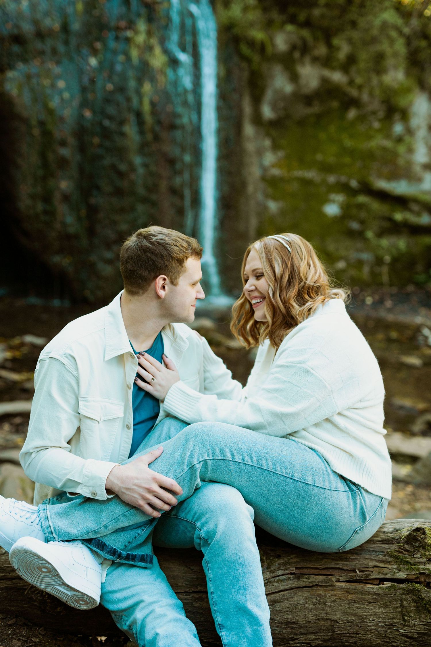 A couple shares an intimate moment while sitting on a fallen log near a waterfall.