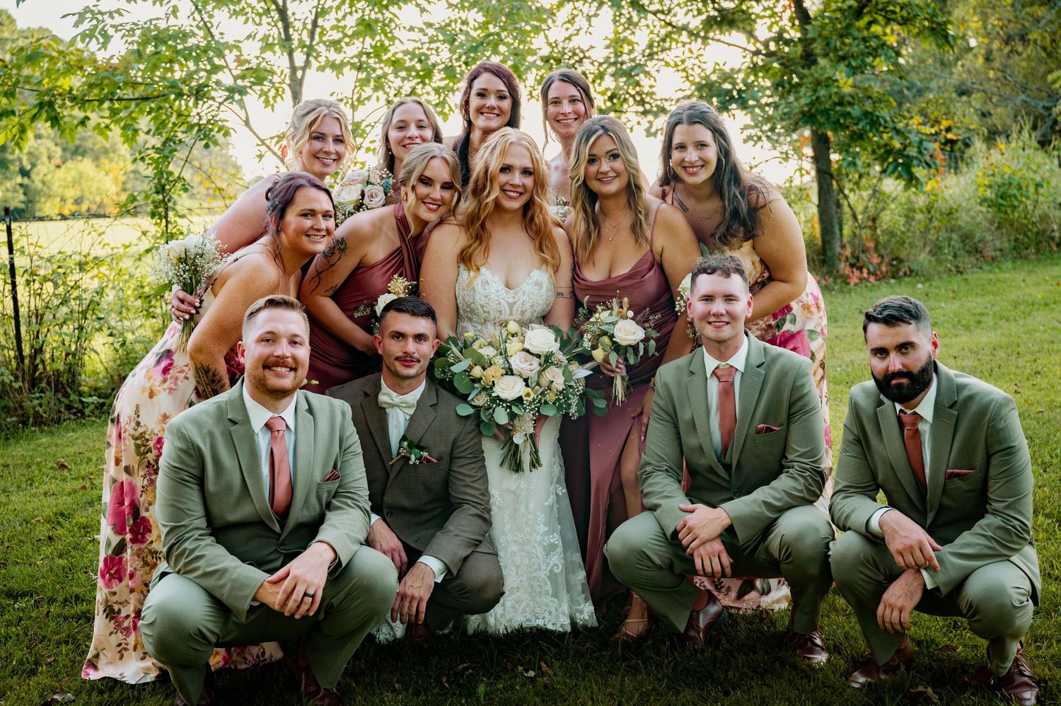 A wedding party poses together outdoors wearing green suits and floral dresses on a sunny summer day.