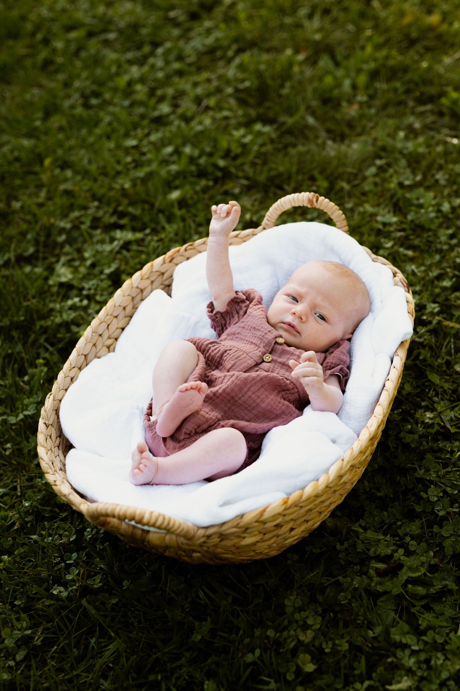 Smiling infant resting in round wicker basket on grassy outdoor setting.