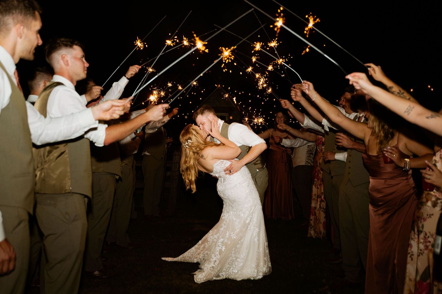 Newlyweds share a romantic kiss under an arch of sparklers held by wedding guests at night.