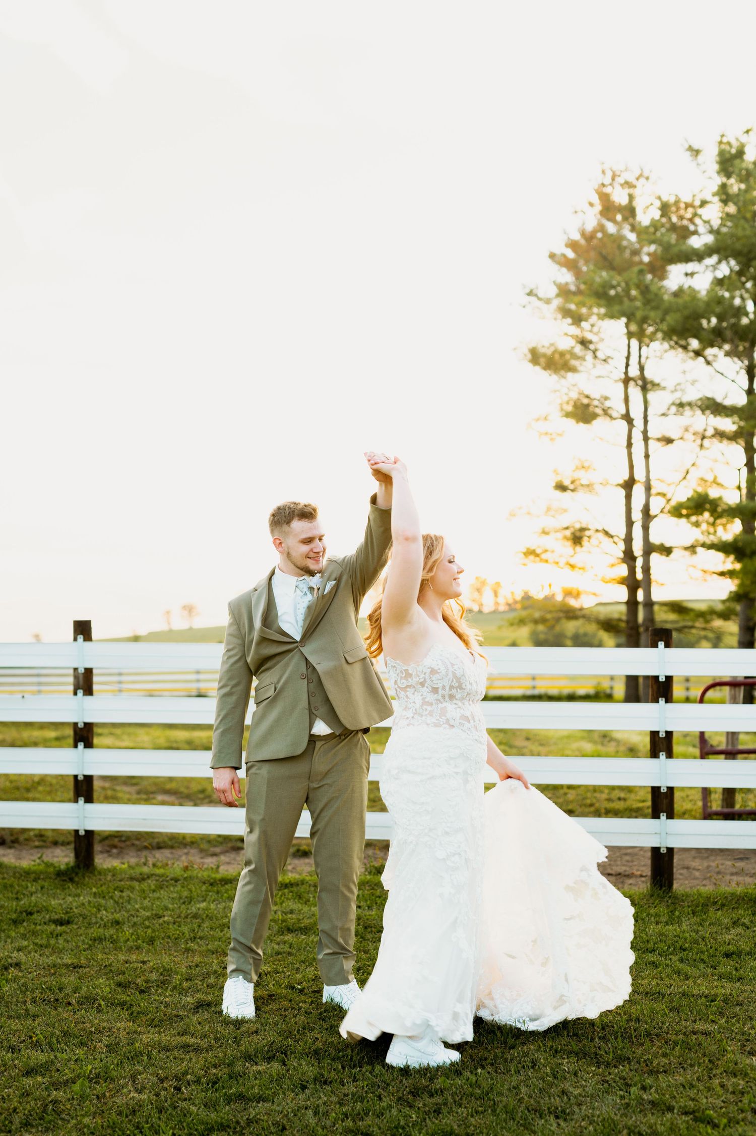 Playful wedding couple dances in an open field with wooden fencing at sunset.
