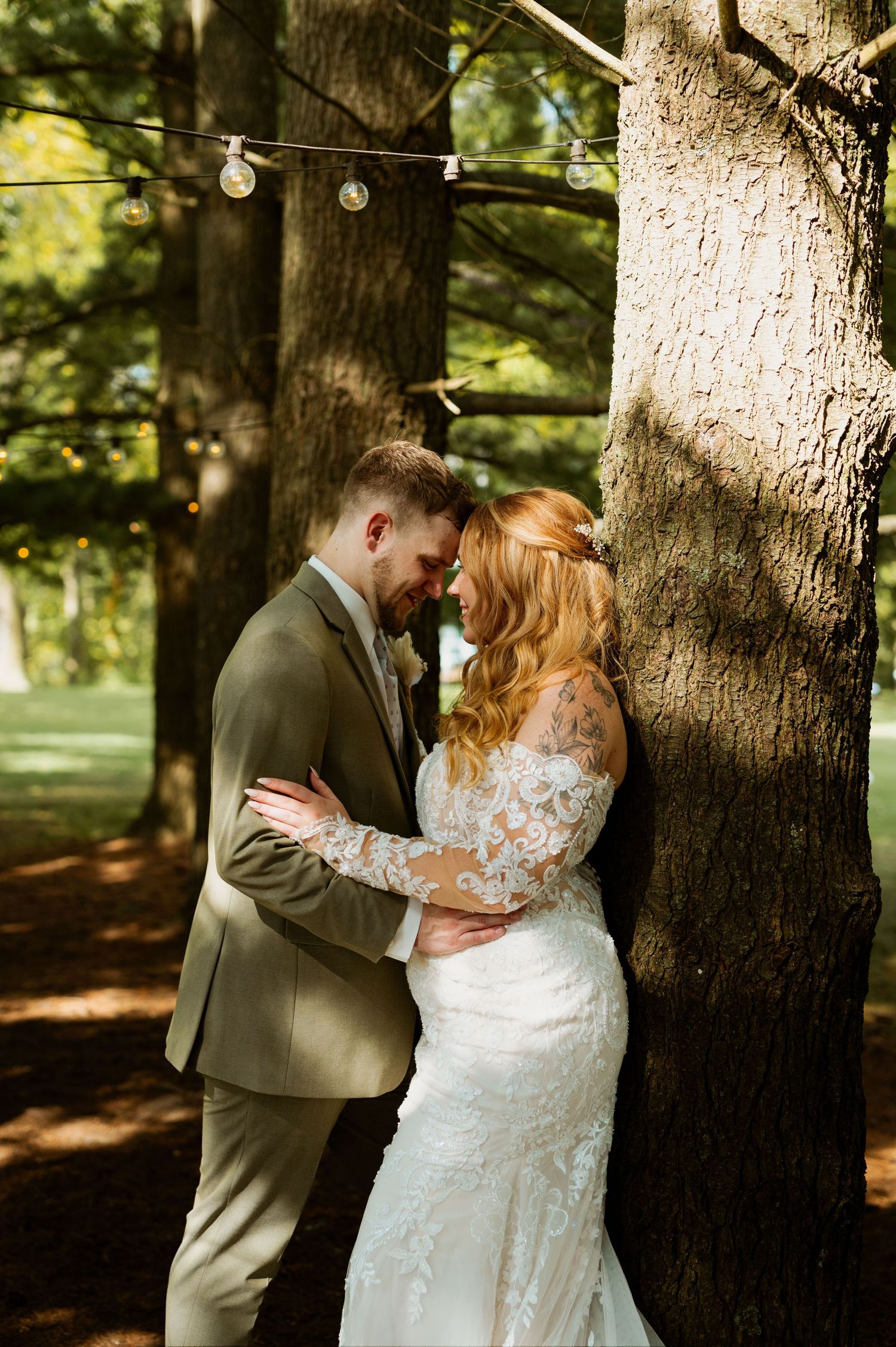 A romantic wedding couple shares intimate moments among tall trees in a wooded setting with string lights overhead.