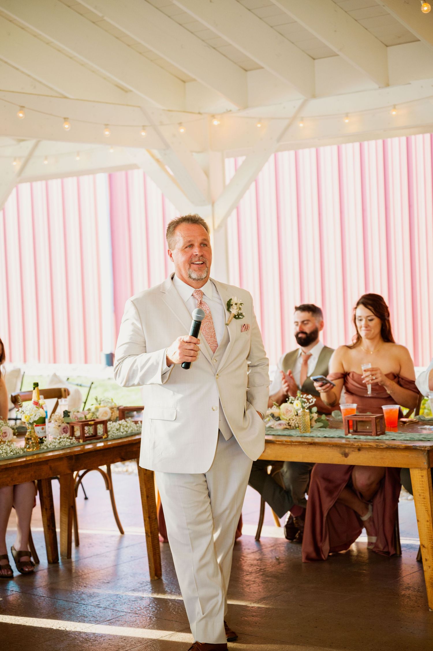 A speaker in a white suit gives a toast during an indoor wedding reception.