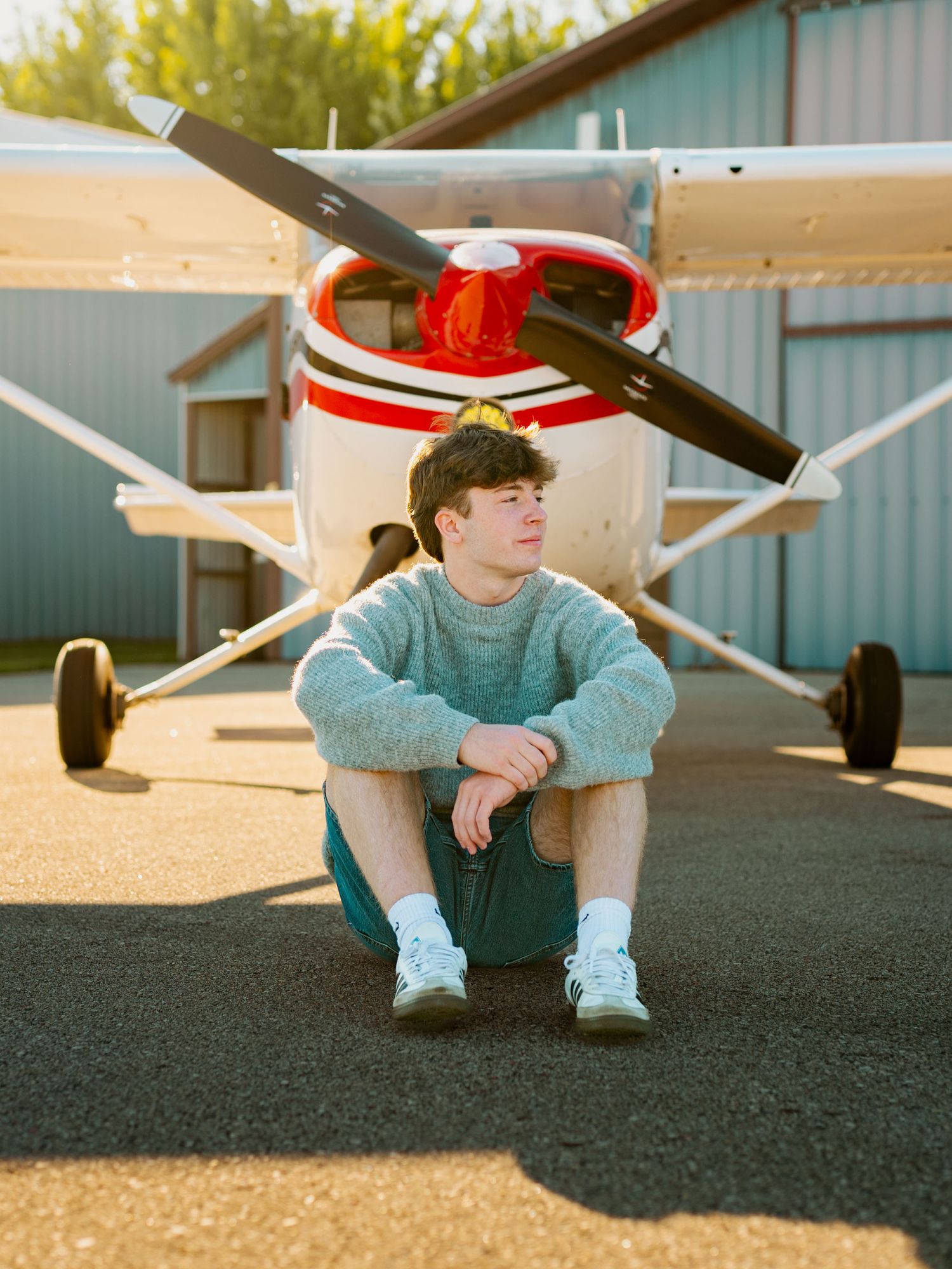 A person sits casually on the ground near a small aircraft on a sunny day at a private airfield.