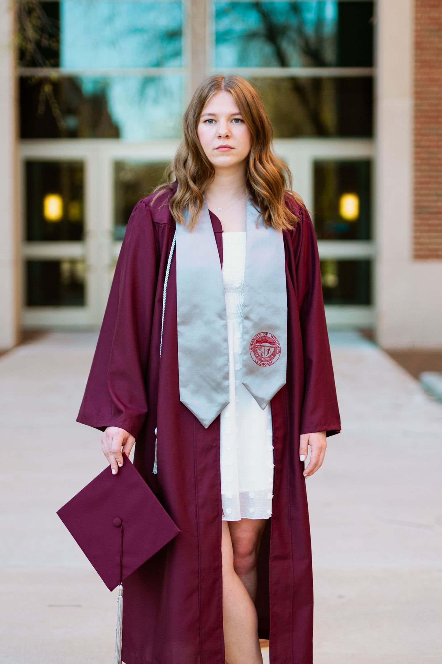 Graduate in maroon cap and gown holding diploma against brick building entrance.