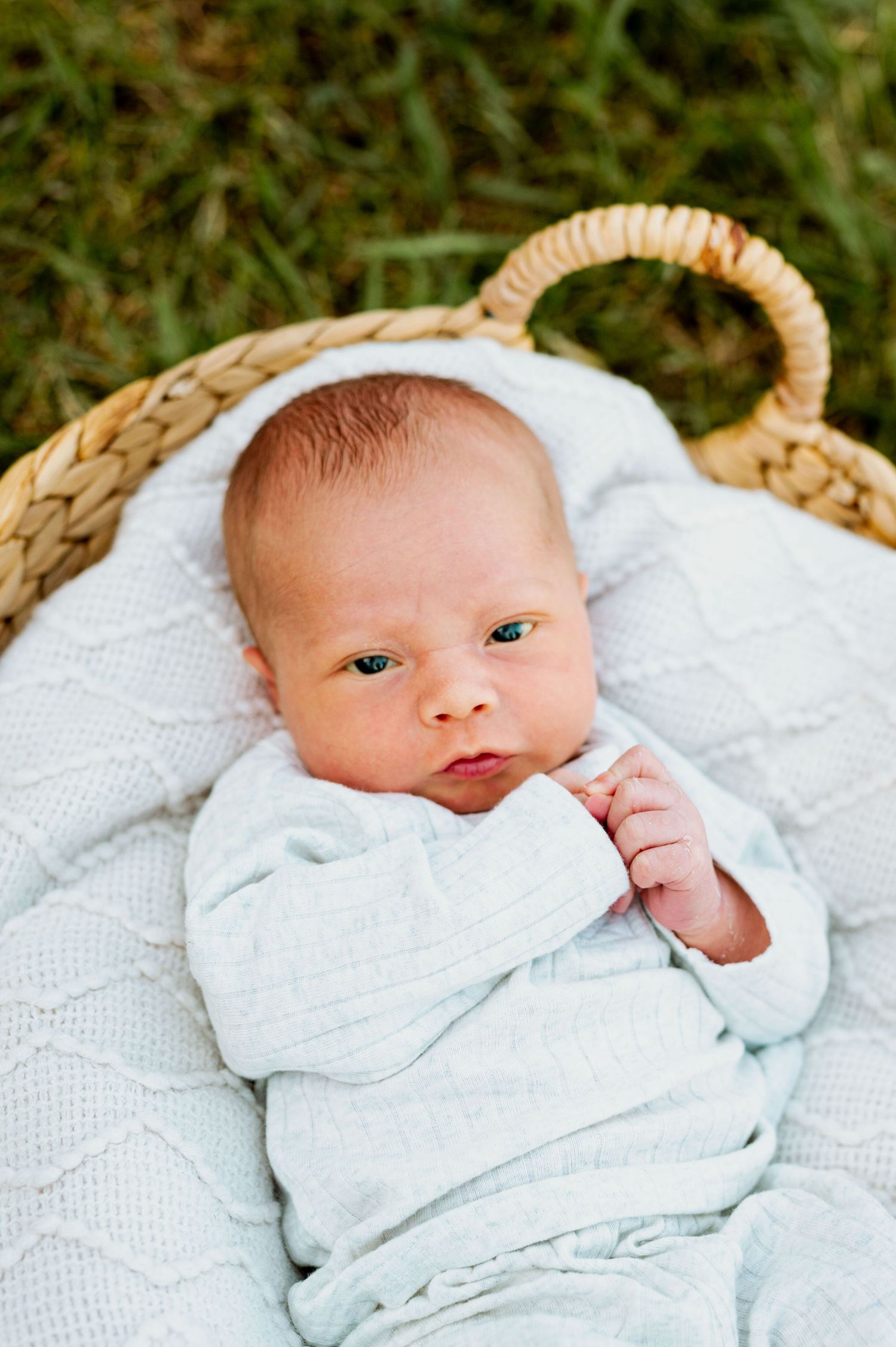 A newborn baby swaddled in white blankets lies on a textured white surface against a green outdoor background.