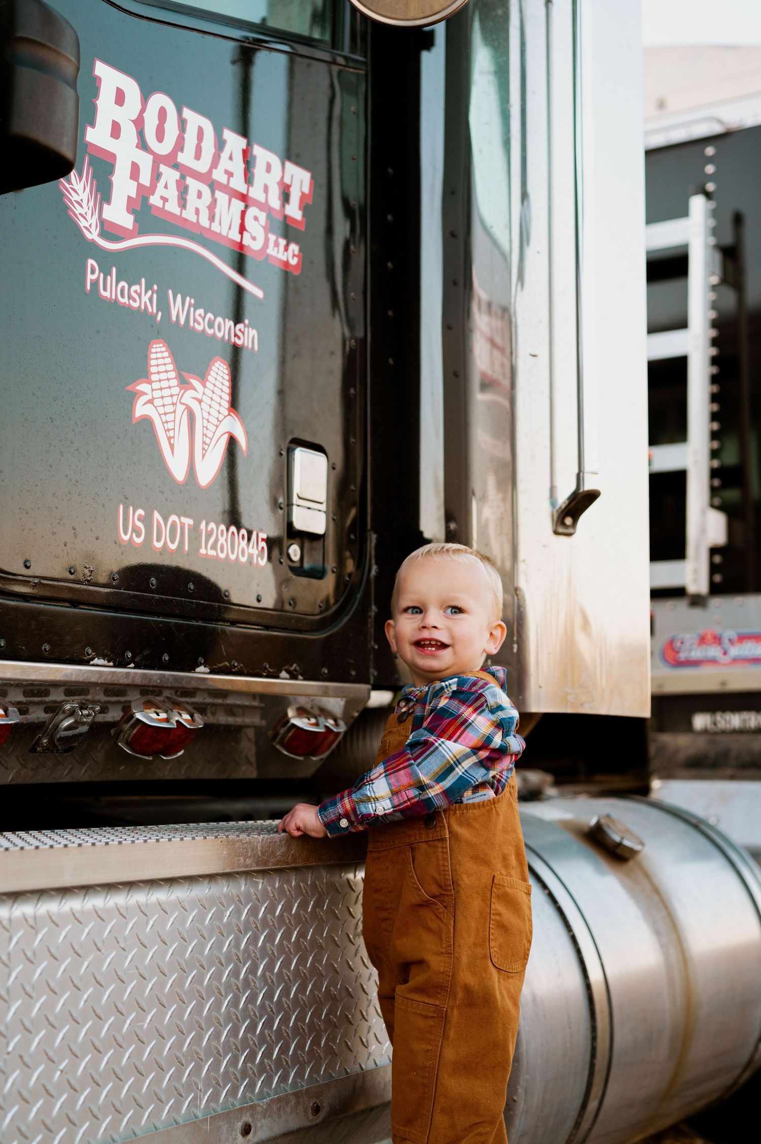 A small child in overalls and plaid shirt sits on the metal step of a red semi truck trailer.