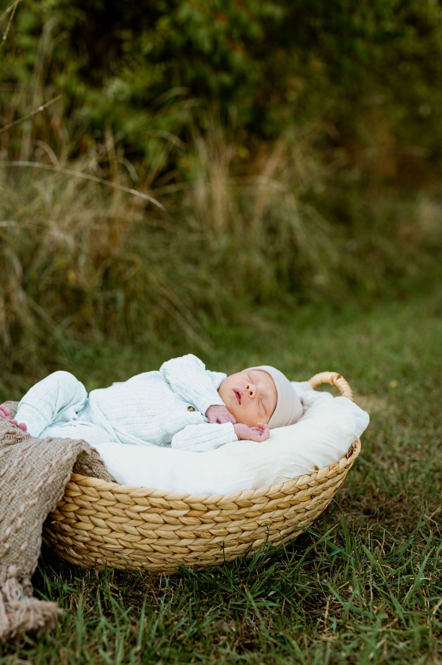 A sleeping newborn baby rests peacefully in a woven basket outdoors on green grass.